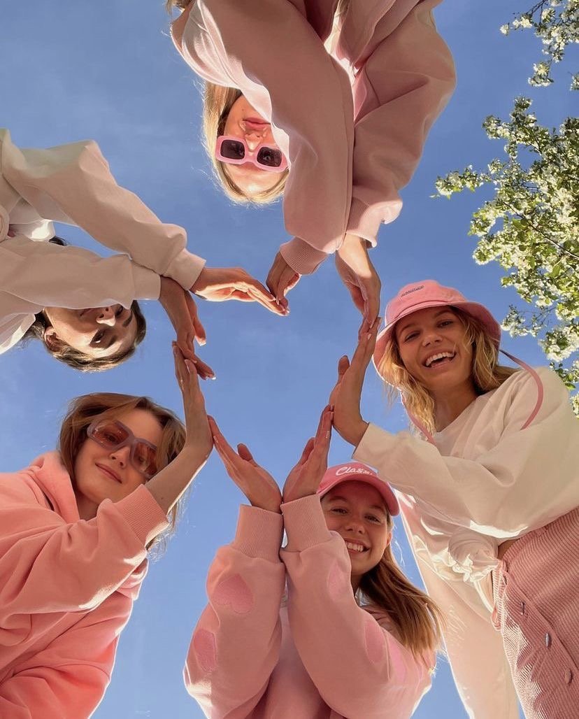 Grupo de jóvenes formando un corazón con las manos desde abajo, contra un cielo azul con árboles en la esquina.