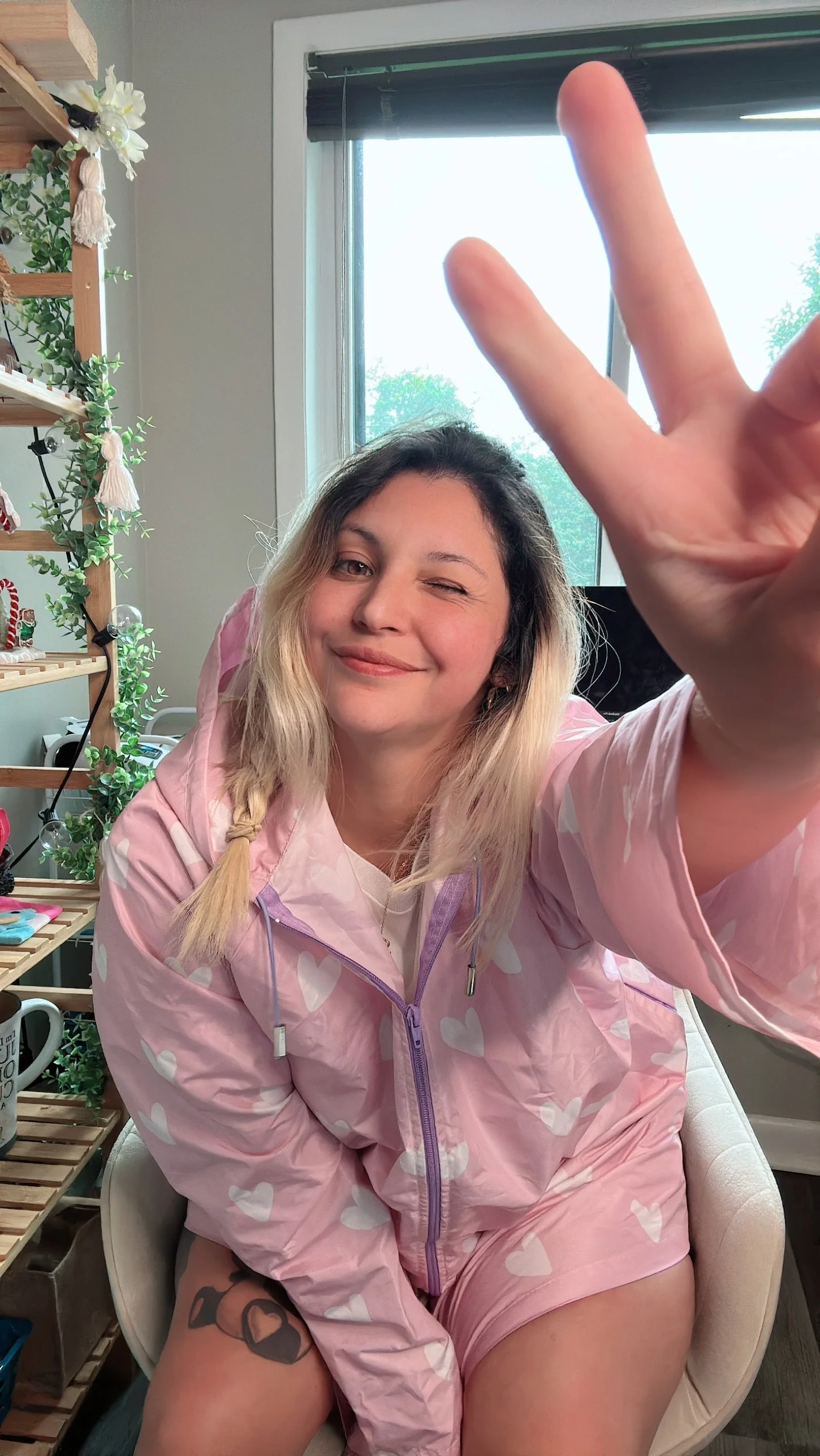 Mujer joven con cabello rubio y ondulado, usando un conjunto rosa con corazones blancos, haciendo una señal de paz con sus dedos y sonriendo en un ambiente interior con decoraciones y una ventana con vista al exterior.