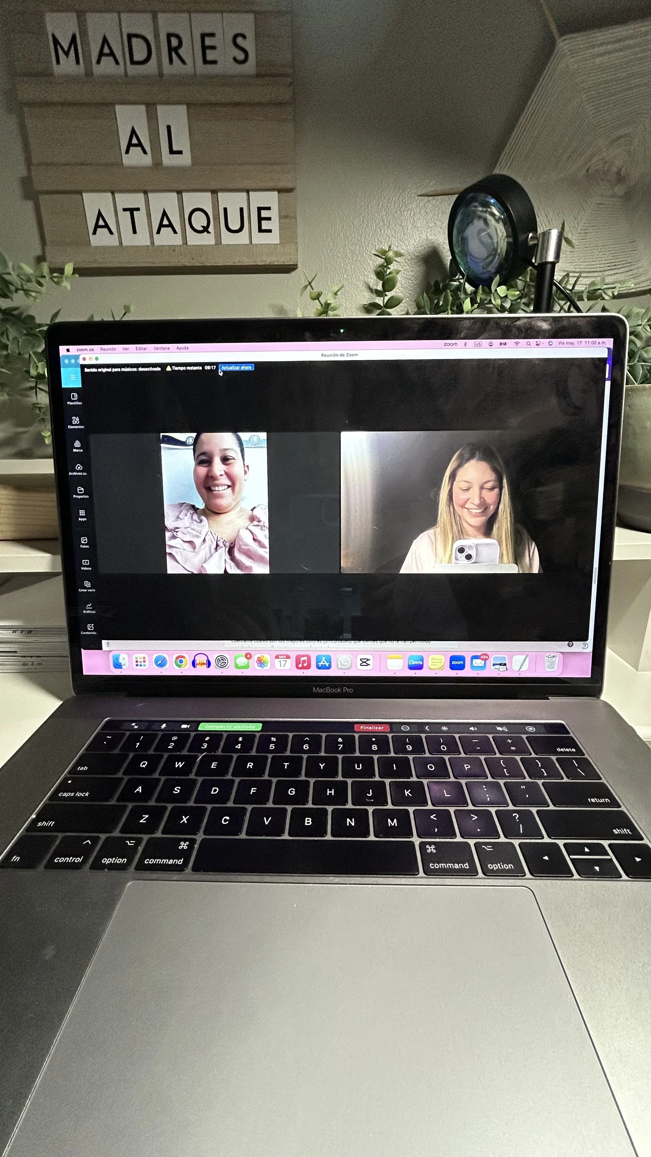 Una computadora portátil MacBook Pro en una mesa con dos mujeres en una videollamada en la pantalla. En el fondo, un cartel que dice "Madres al Ataque" en letras mayúsculas en una pizarra de madera.