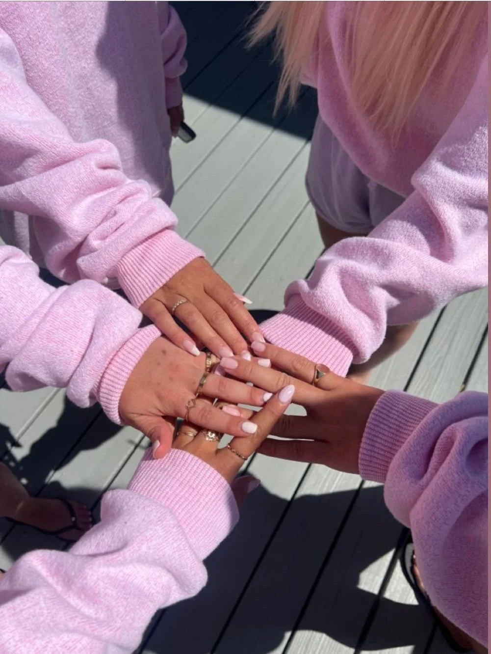 Varias manos con diferentes tonos de piel y anillos emparejadas en el centro, sobre una superficie de madera, todas usando sudaderas rosadas.