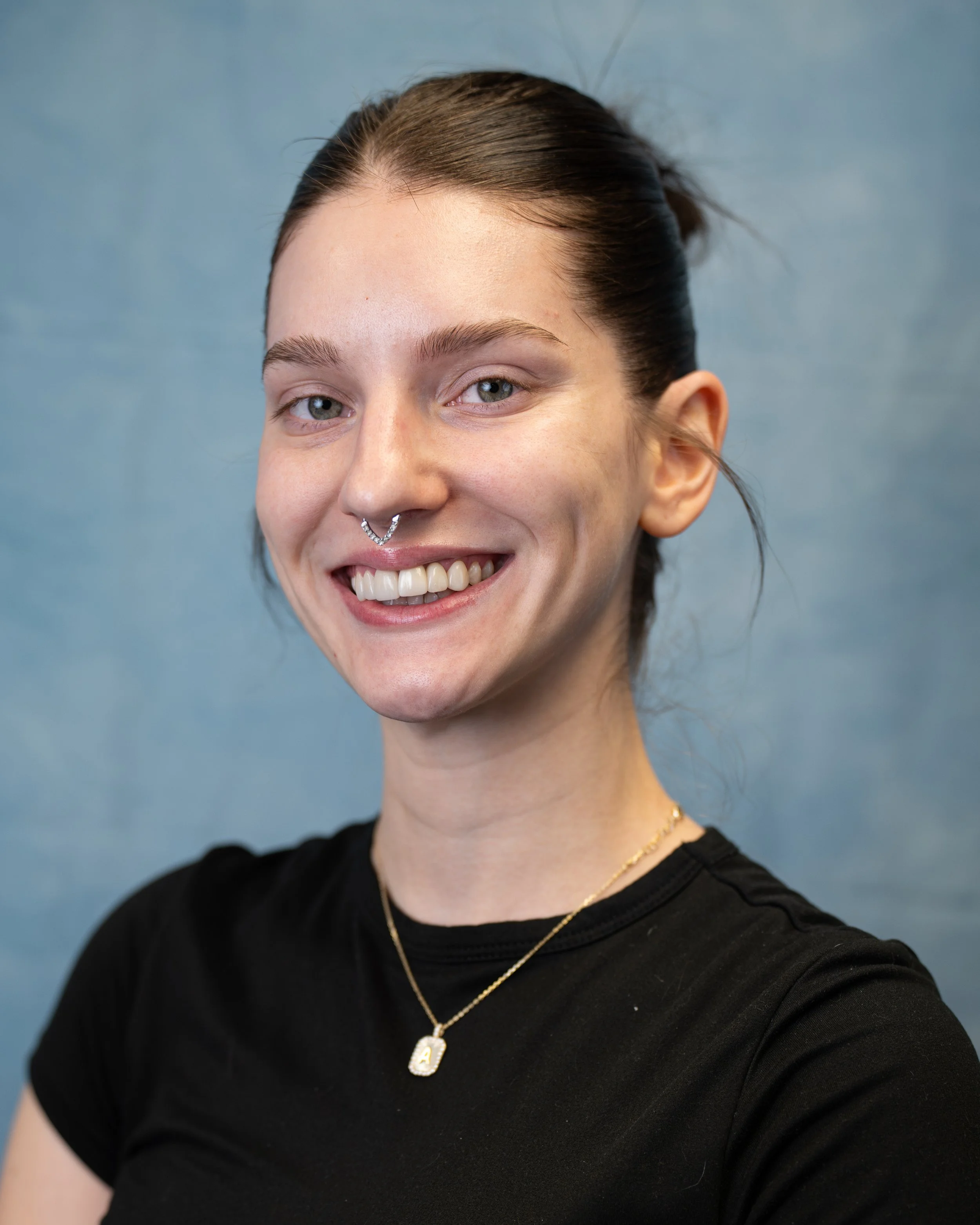 A young woman with brown hair tied back, wearing a septum piercing, a black shirt, and gold jewelry, smiling at the camera against a blue background.