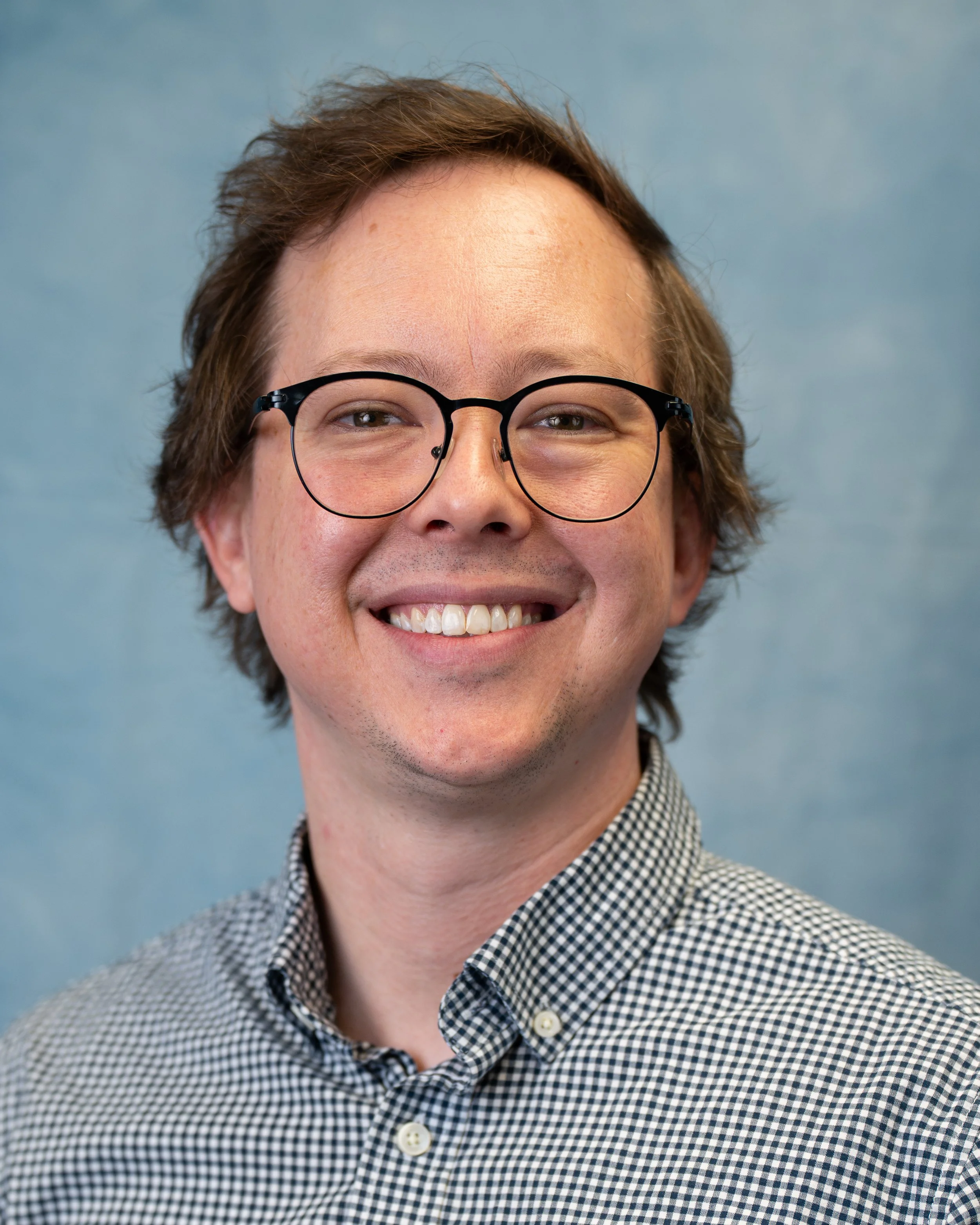 Portrait of a young man with brown hair, glasses, and a checkered shirt, smiling against a blue background.