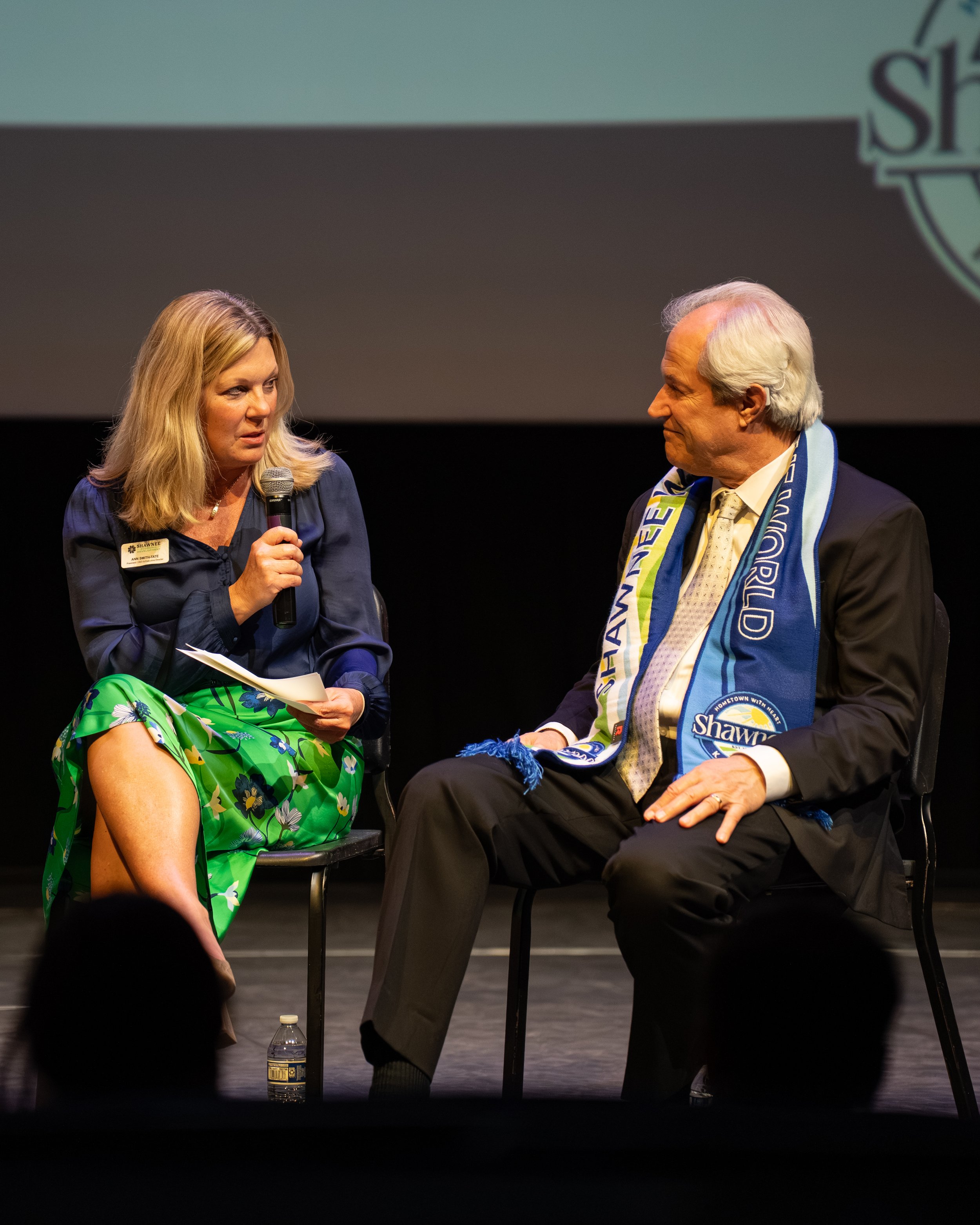 A woman holding a microphone and speaking to a man with a Shawnee County Democratic scarf on stage during an event.