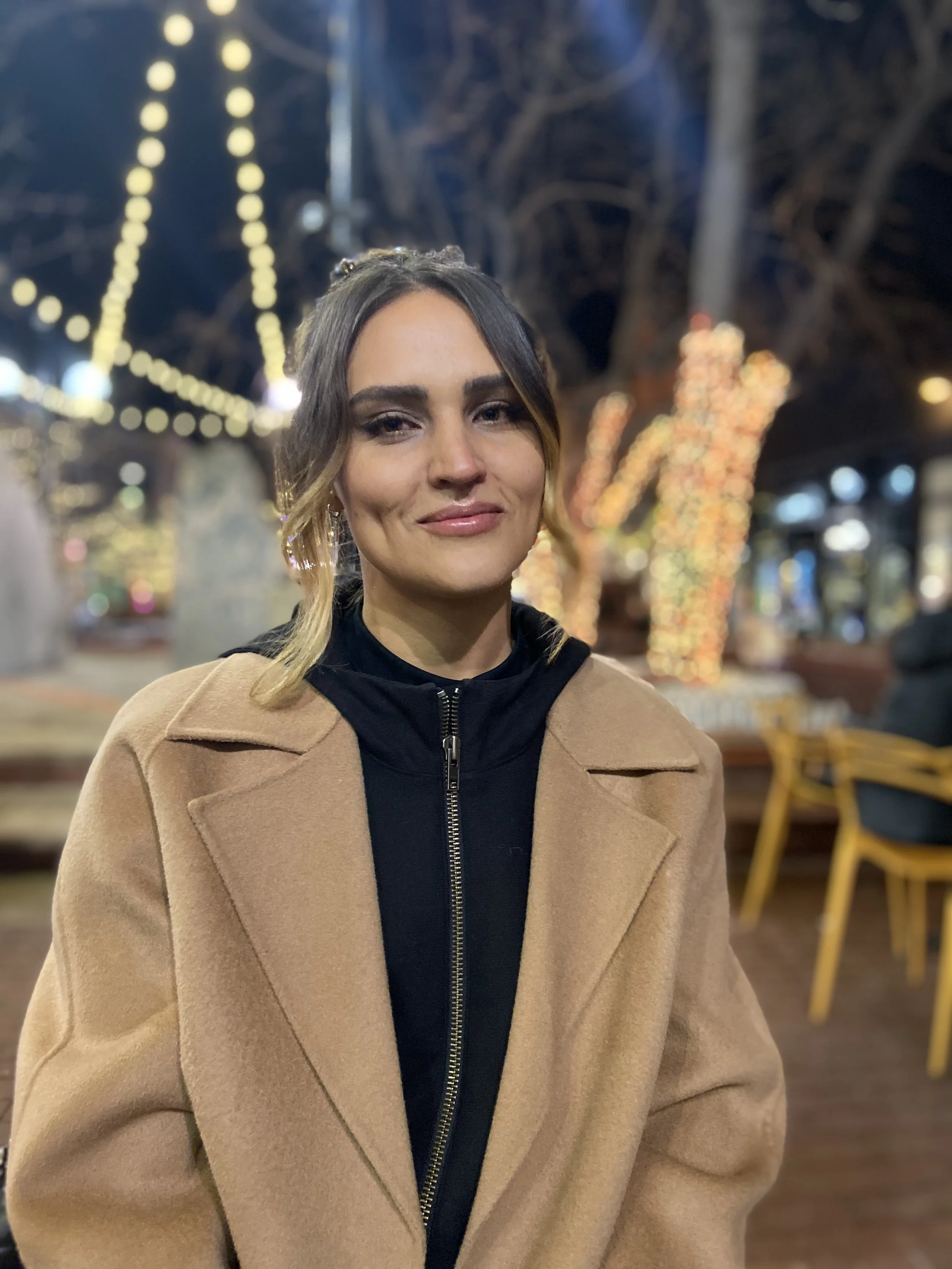 A woman with light skin and brown hair posed outdoors at night, wearing a tan coat over a black top, with blurred festive lights and trees in the background.