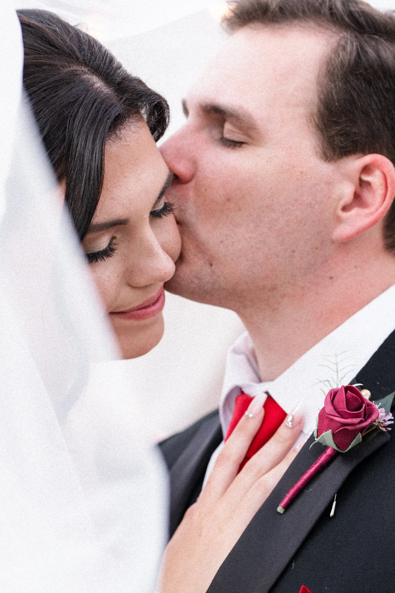 A close-up of a DFW newlywed couple kissing, with the bride wearing makeup and the groom in a tuxedo with a red boutonniere.