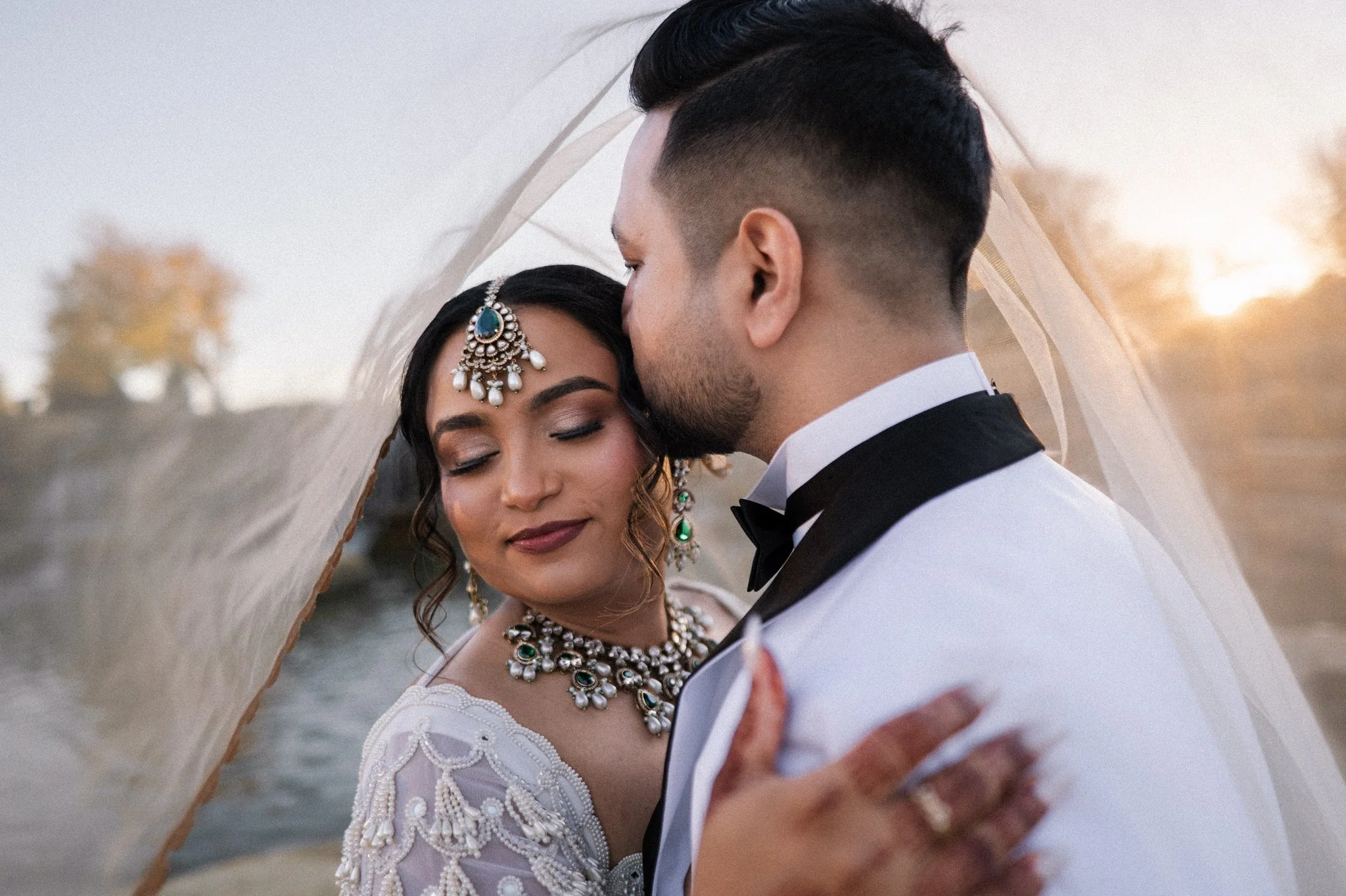 Bride and groom embracing under bride's veil. Groom kissing side of bride's head.