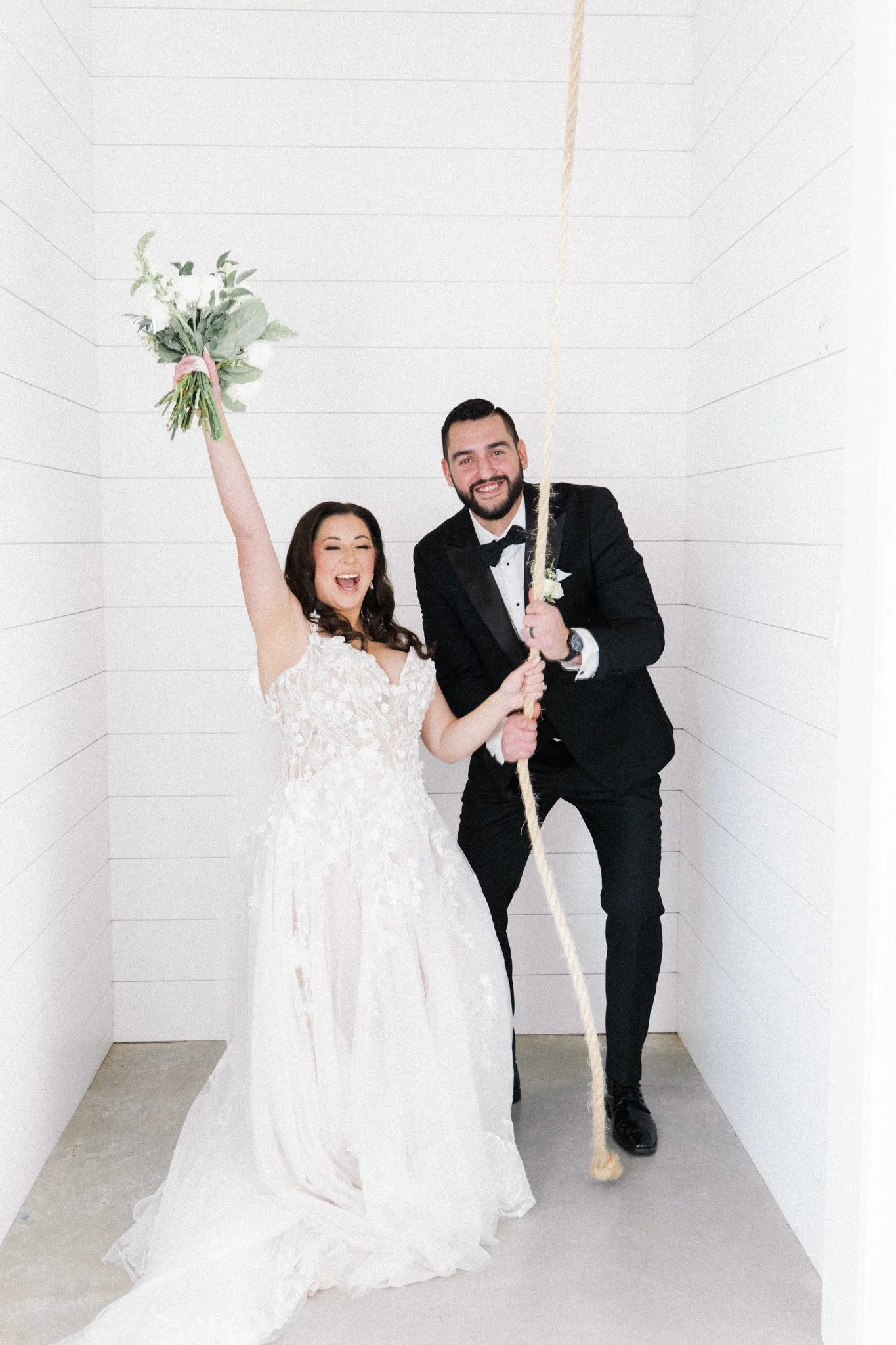 Bridge and groom holding a wedding bell rope. Bride has one arm up in the air holding bouquet and smiling.
