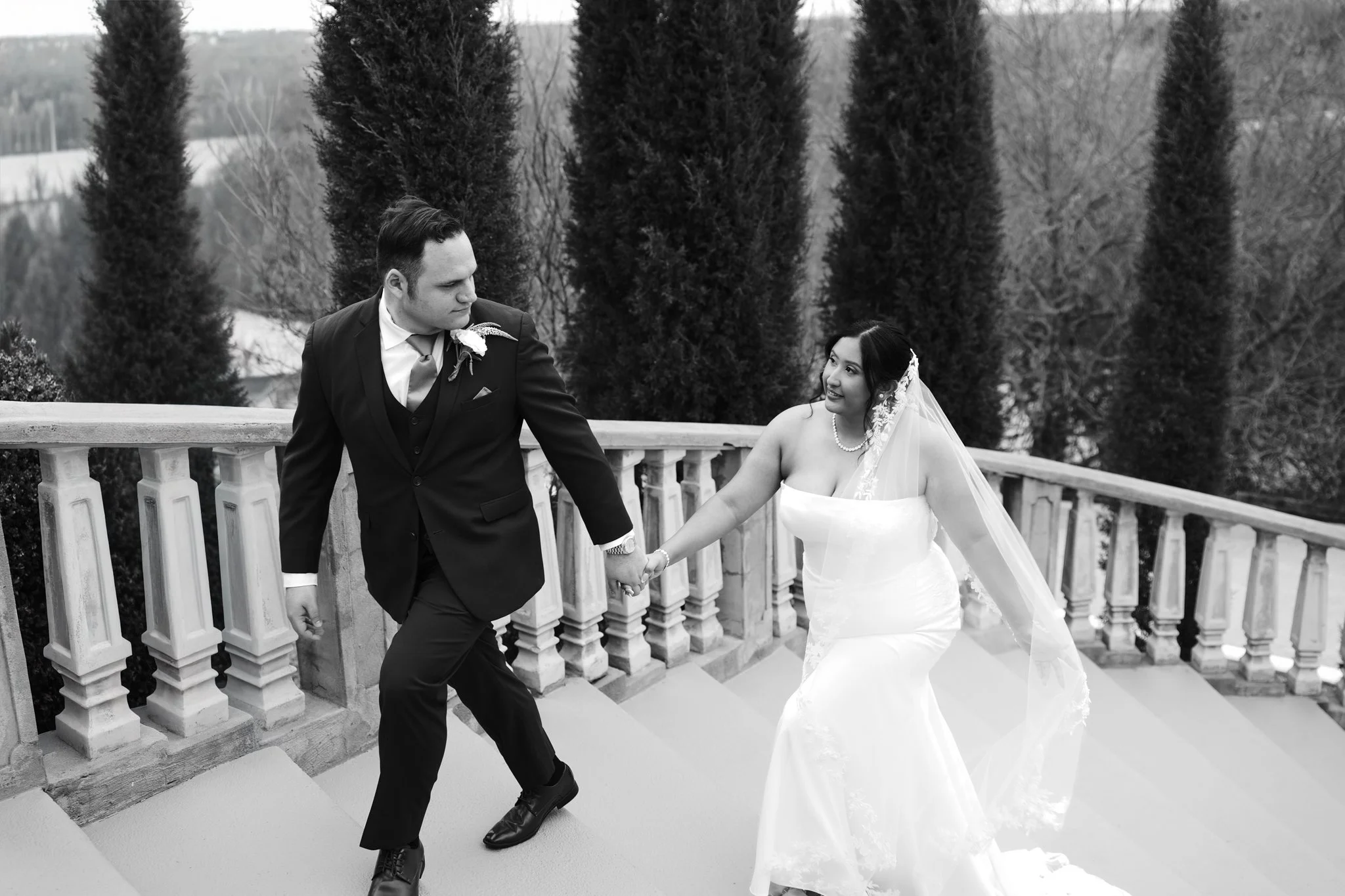 A black and white photo of a DFW bride and groom dancing outdoors on a balcony with a stone railing, surrounded by tall trees in a winter landscape.