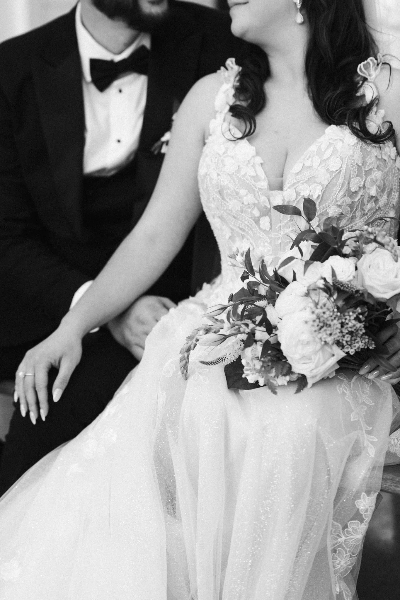 A Dallas bride and groom sitting close together, with the bride holding a bouquet of flowers, wearing a lace wedding dress, and the groom in a tuxedo with a bow tie, captured in a black and white photograph.