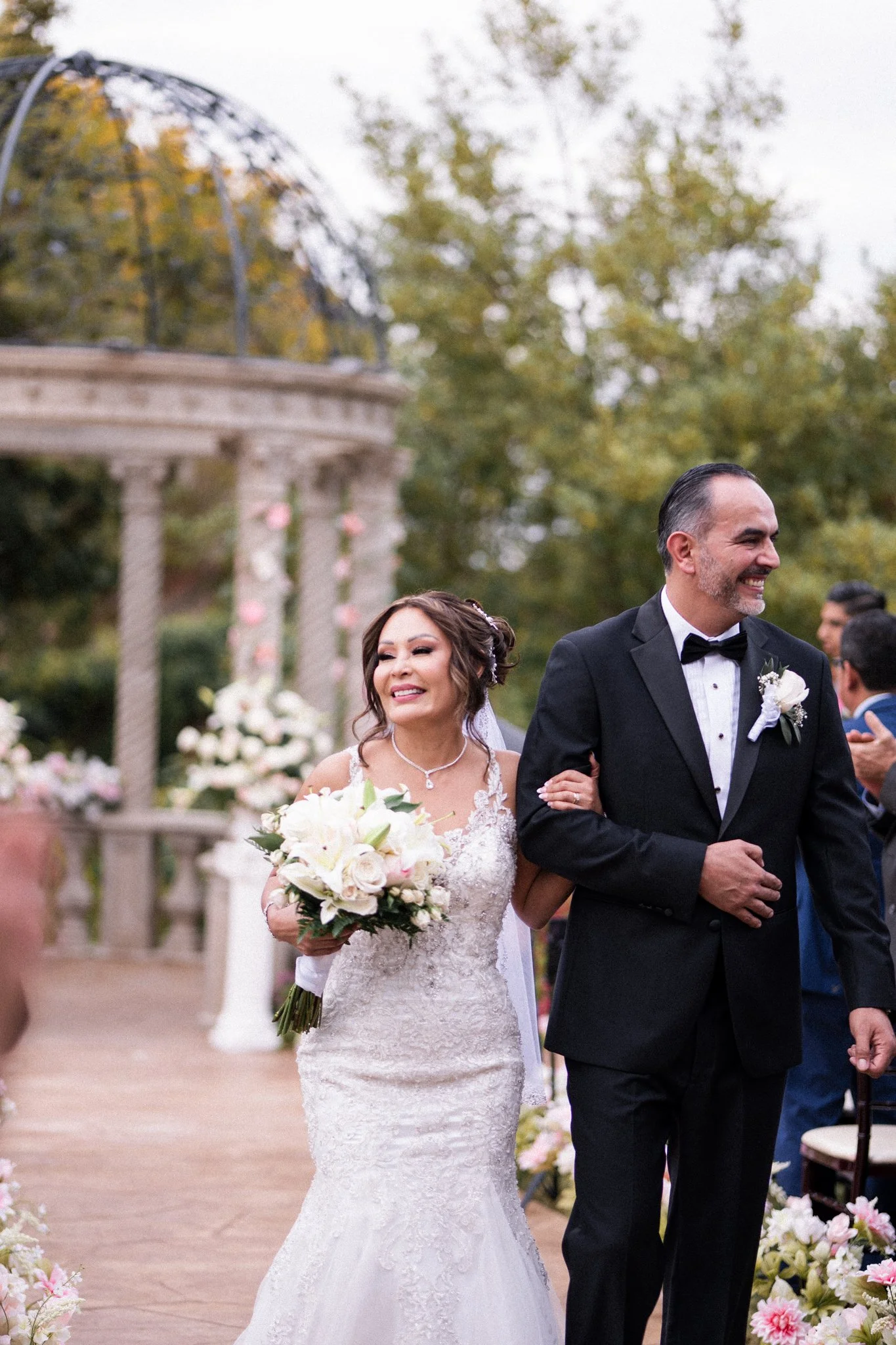 A DFW bride and groom smiling and standing arm in arm during a wedding ceremony outdoors with floral decorations and trees in the background.