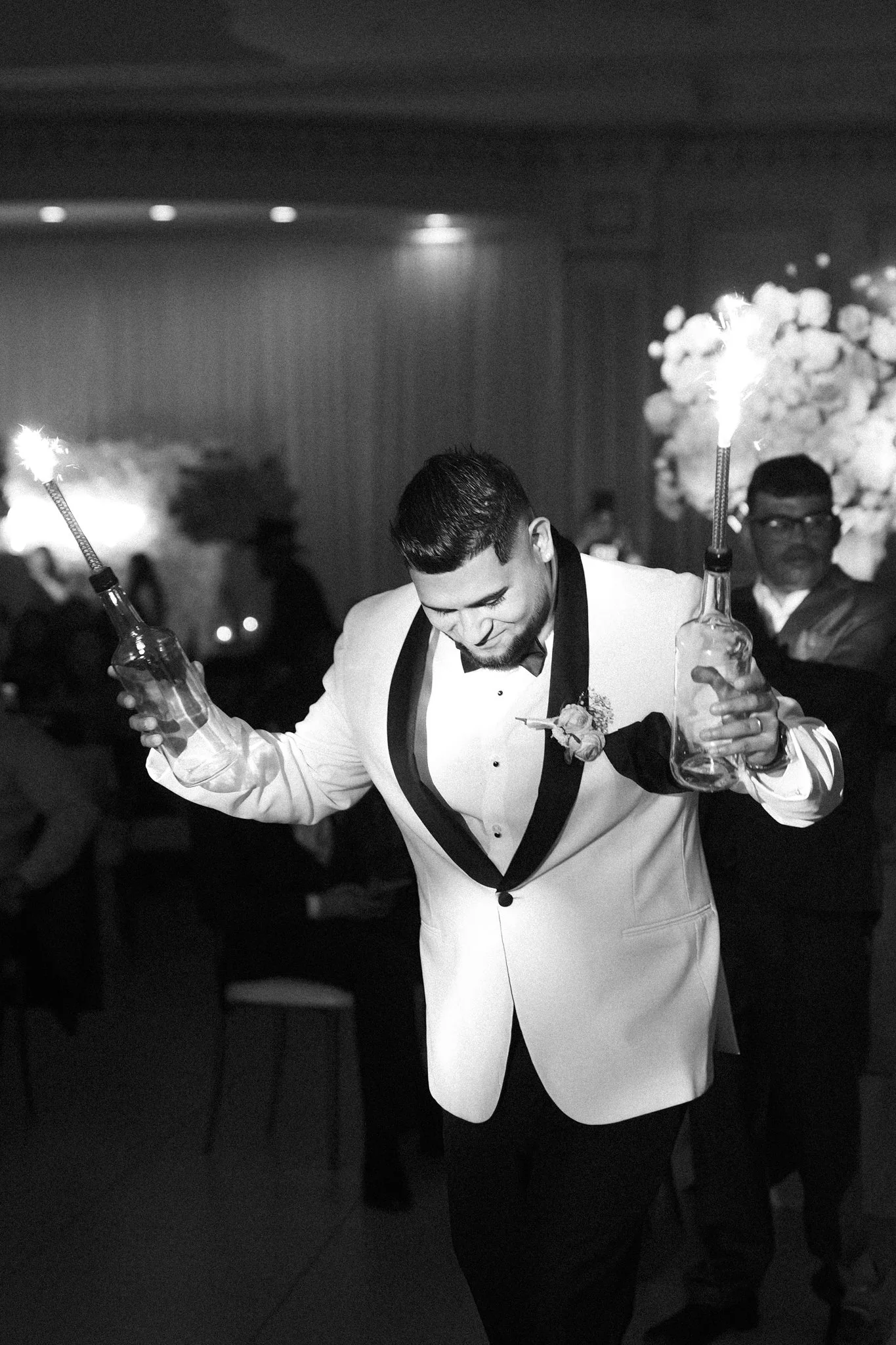 A DFW groom in a tuxedo with a boutonnière, holding two bottles with sparklers attached, at a celebration or special event. He is smiling and appears to be enjoying the moment.