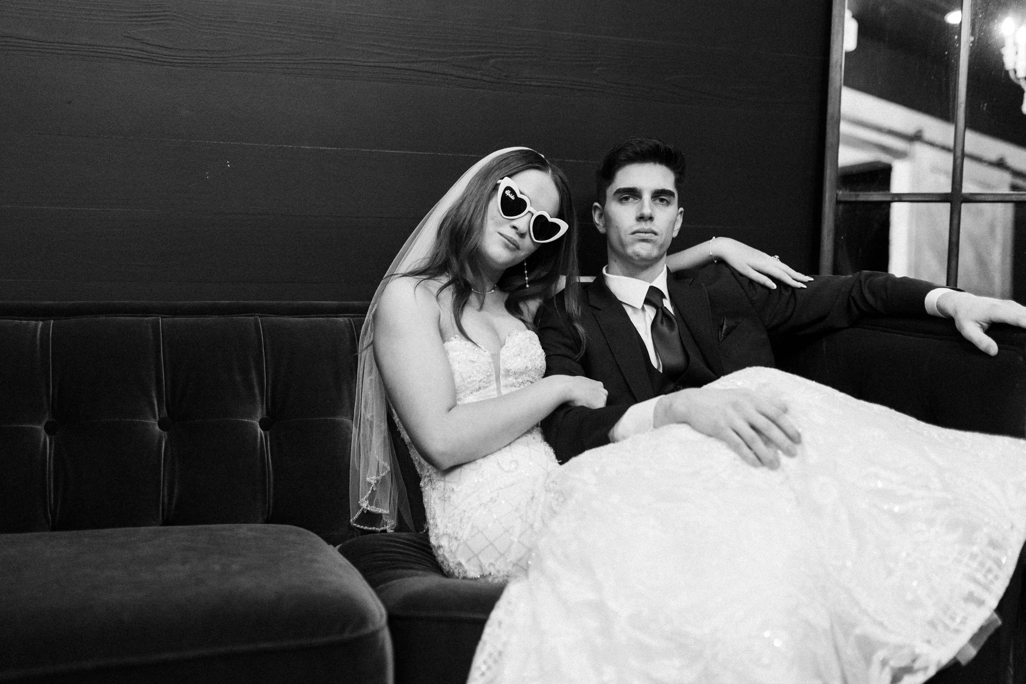 A black-and-white photo of a DFW bride and groom sitting on a bench, with the bride wearing heart-shaped sunglasses and the groom dressed in a suit and tie, both looking at the camera.