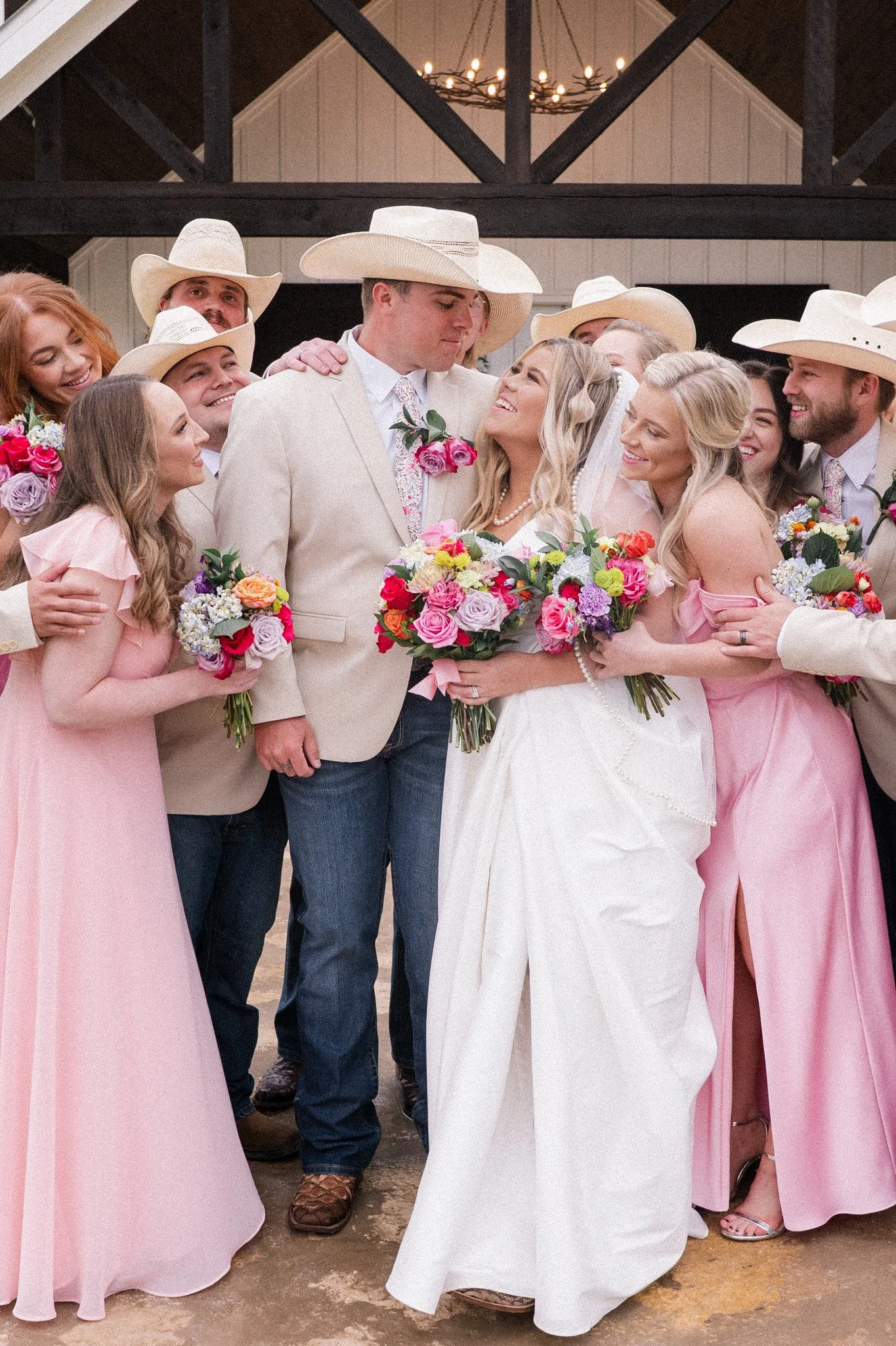A wedding reception scene featuring a bride and groom surrounded by friends or family members. The bride is wearing a white dress and holding a bouquet, while the groom is in a light-colored suit with a pink boutonniere. Several women are dressed in pink dresses with bouquets, and men are wearing white shirts, pink ties, and cowboy hats. The setting appears to be indoors with rustic decor and a chandelier above.