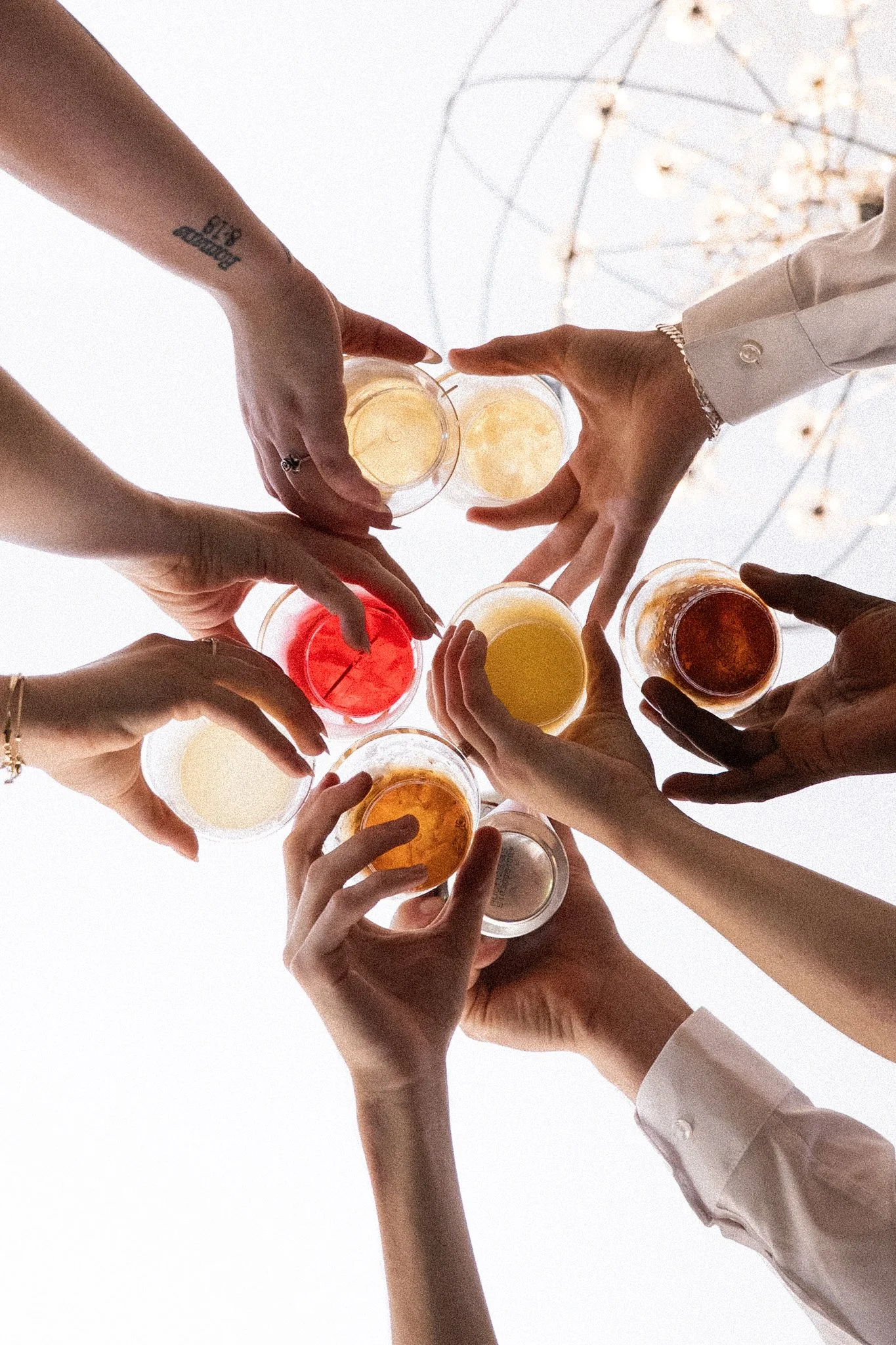 Multiple wedding guests raising glasses for a toast, viewed from below, with a bright background and a chandelier