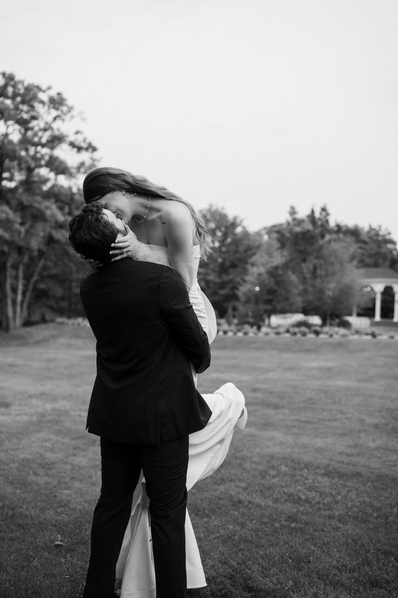 A black and white photo of a DFW newlywed couple outdoors, one person lifting the other in a kiss, with trees and a field in the background.