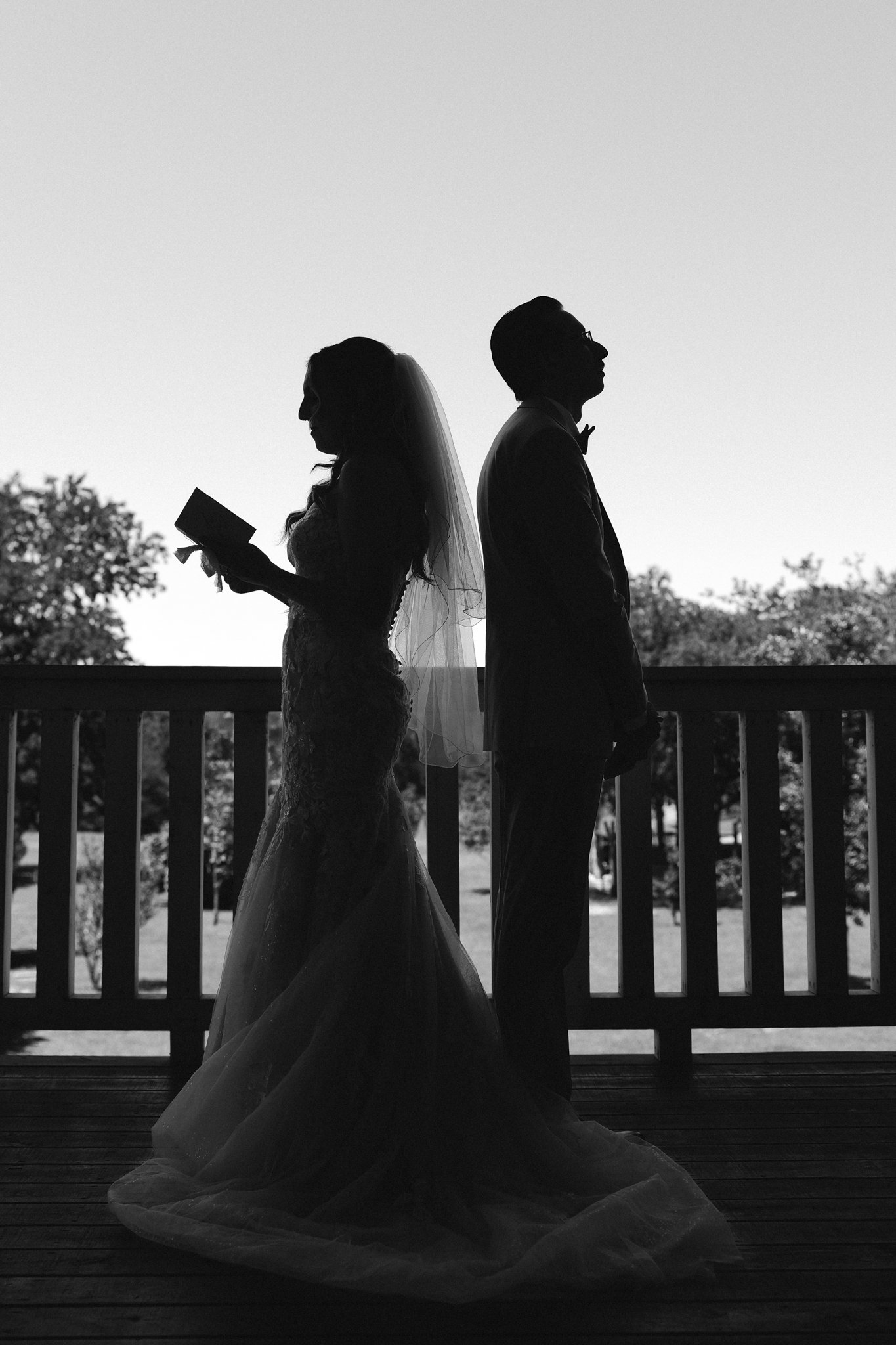 Black and white silhouette of a Dallas bride and groom standing back to back on a wooden deck with a railing, outdoors with trees in the background, during sunset or sunrise.