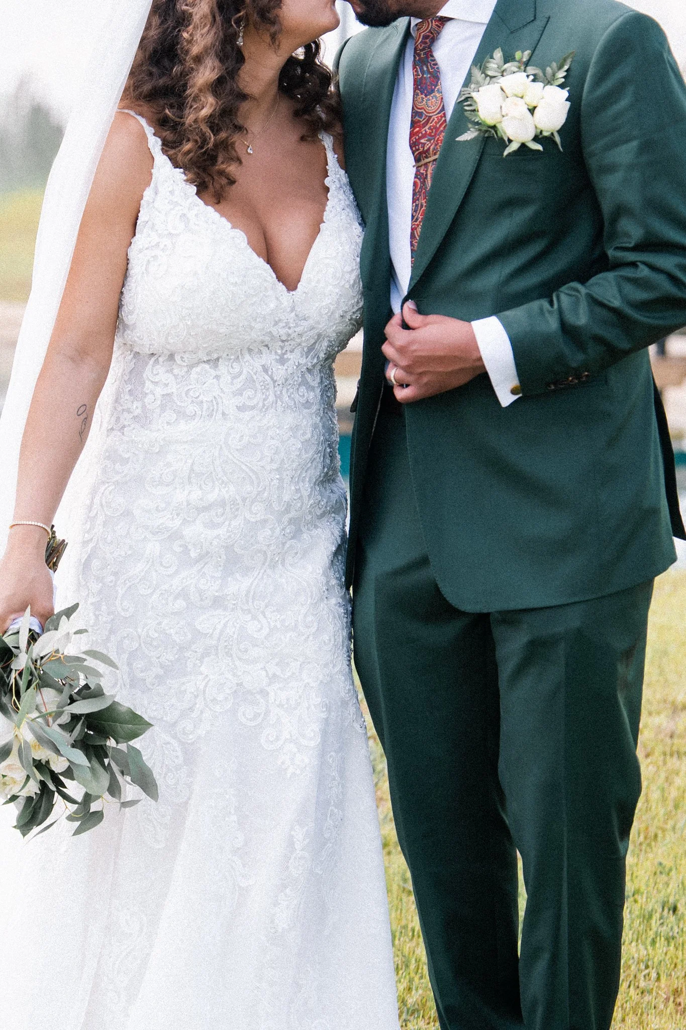 A bride and groom standing close together at their wedding, with the bride wearing a white lace wedding dress and holding a bouquet of greenery, and the groom dressed in a green suit with a white shirt, colorful tie, and a boutonniere of white flowers.