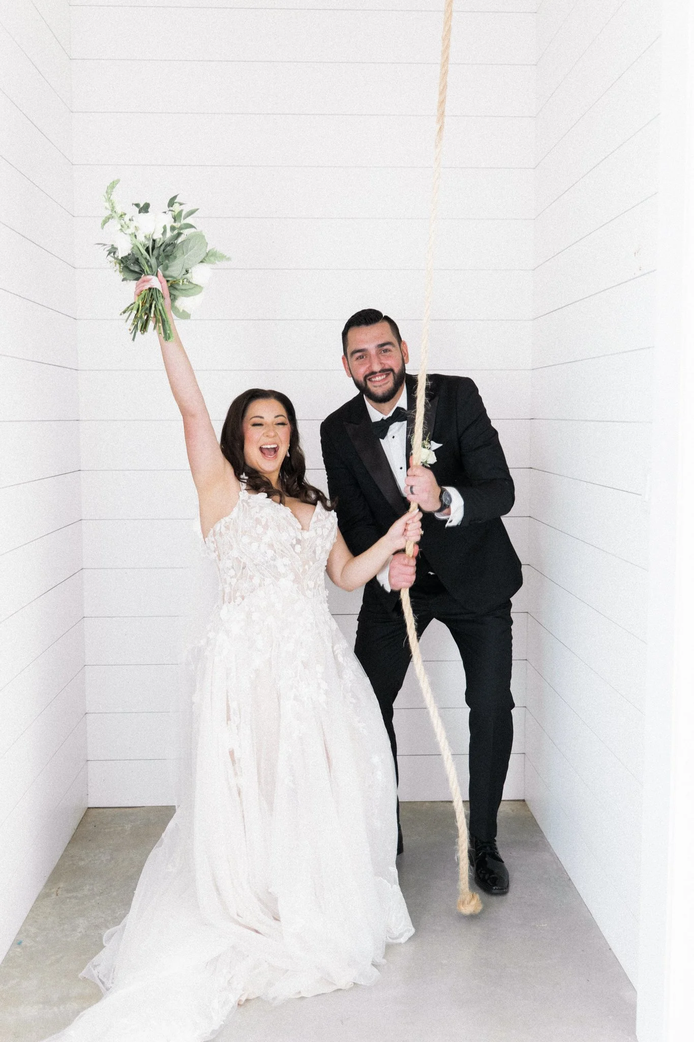 A Dallas newlywed couple celebrating, with the bride holding a bouquet and the groom pulling a rope, inside a white room with horizontal paneling.