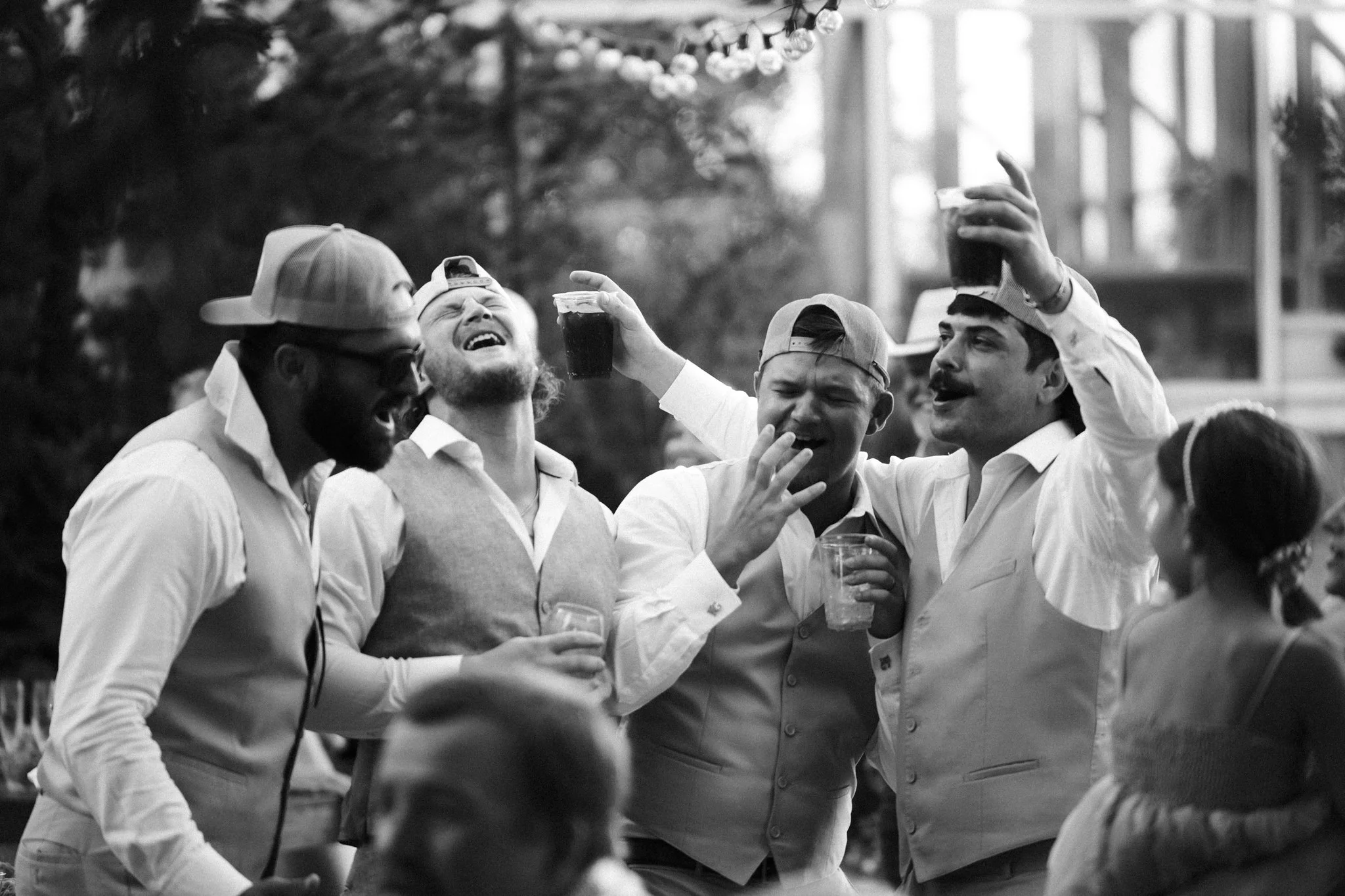 A black and white photo of five men enjoying a drink at an outdoor gathering. They are dressed in light-colored shirts and are wearing baseball caps. Some are laughing, smiling, and holding drinks, with a festive atmosphere suggested by blurred string lights in the background.