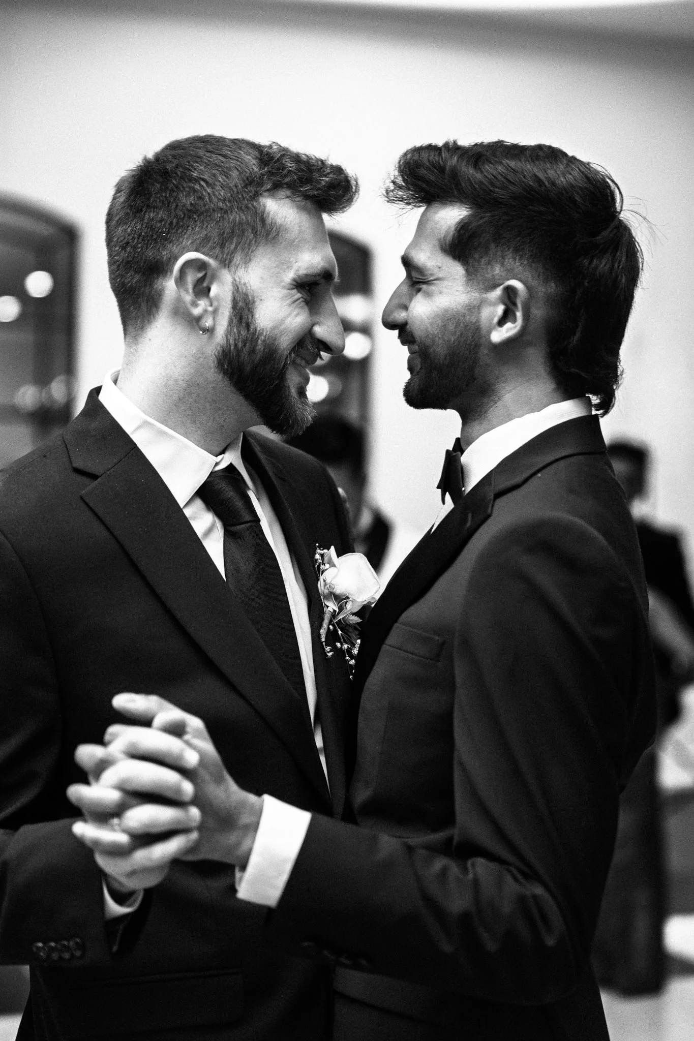 Two men dressed in tuxedos are facing each other closely, smiling, with their foreheads touching, at what appears to be a wedding or formal event.