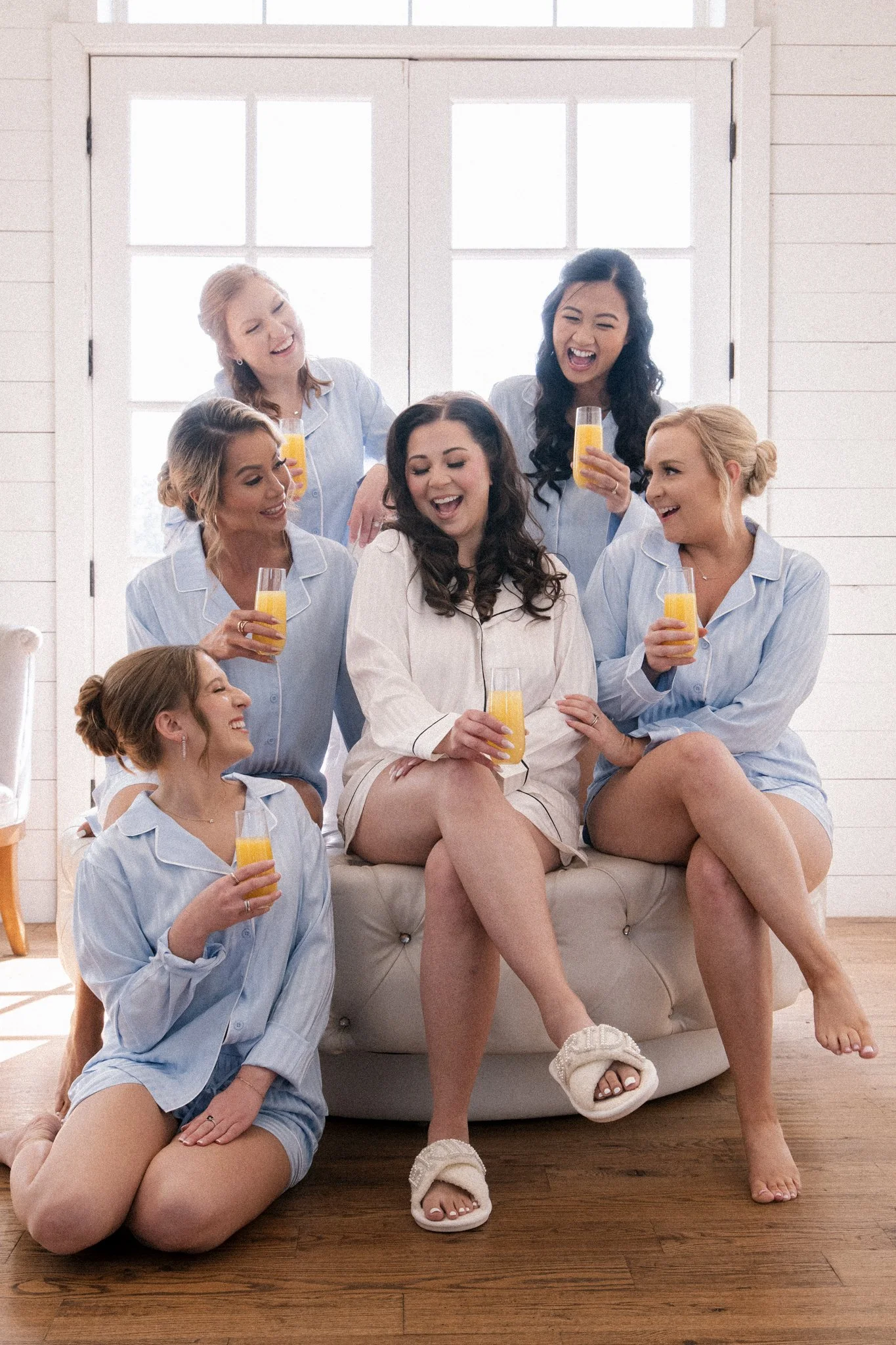 A DFW bride and five bridesmaids wearing matching light blue pajamas gathered in a bright room, with some sitting on a sofa and others standing, holding glasses of orange juice, laughing and enjoying each other's company.