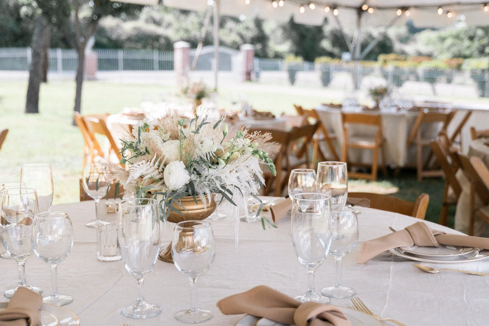 A table set for a formal outdoor wedding reception with glassware, napkins, and a centerpiece of white flowers and greenery, with a canopy and string lights above and a fenced yard in the background.