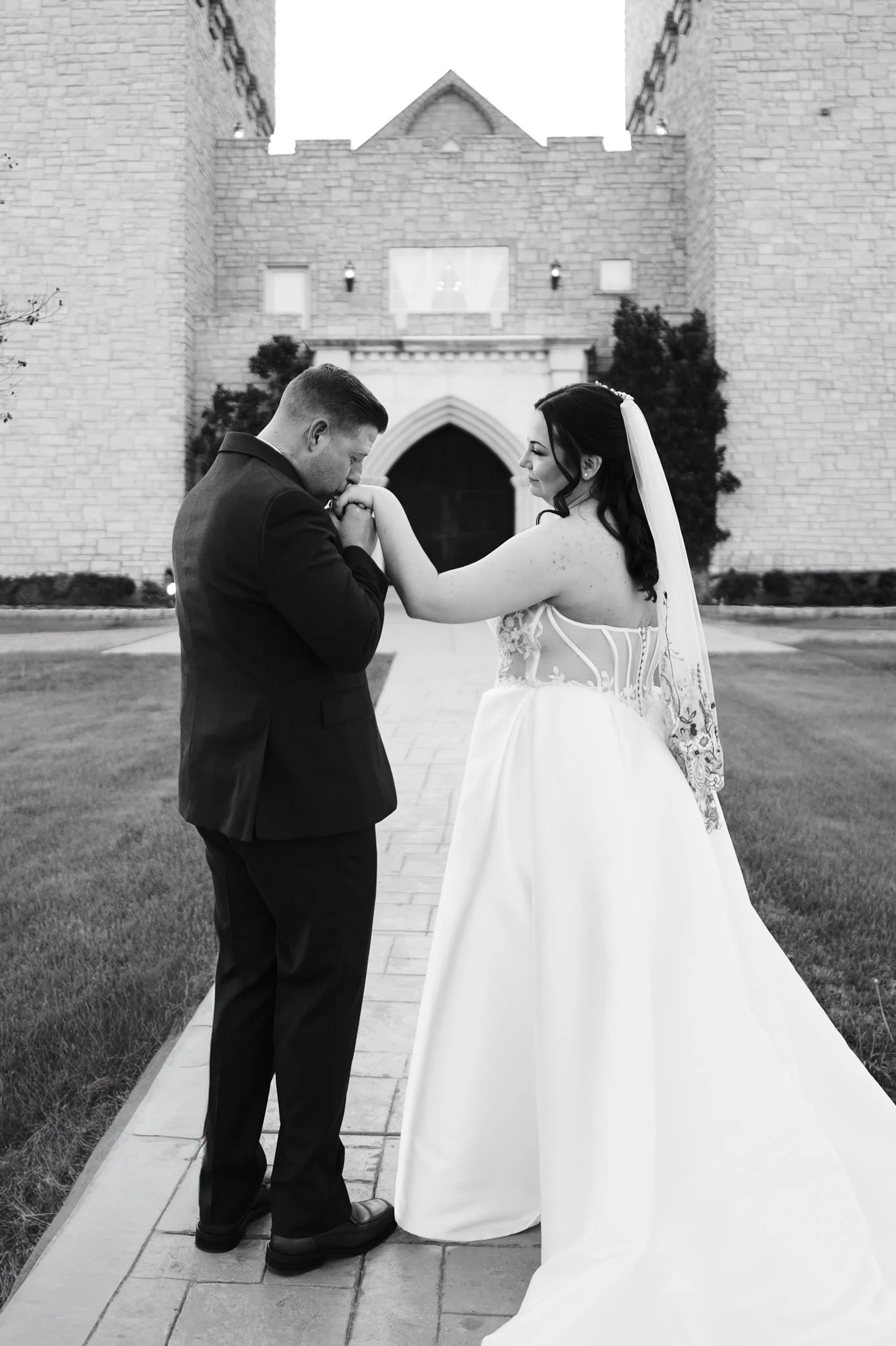 Black and white photo of a Dallas bride and groom outside a brick building. The groom is kissing the bride's hand, and they are smiling at each other. The bride is wearing a white wedding dress with a veil, and the groom is in a dark suit.