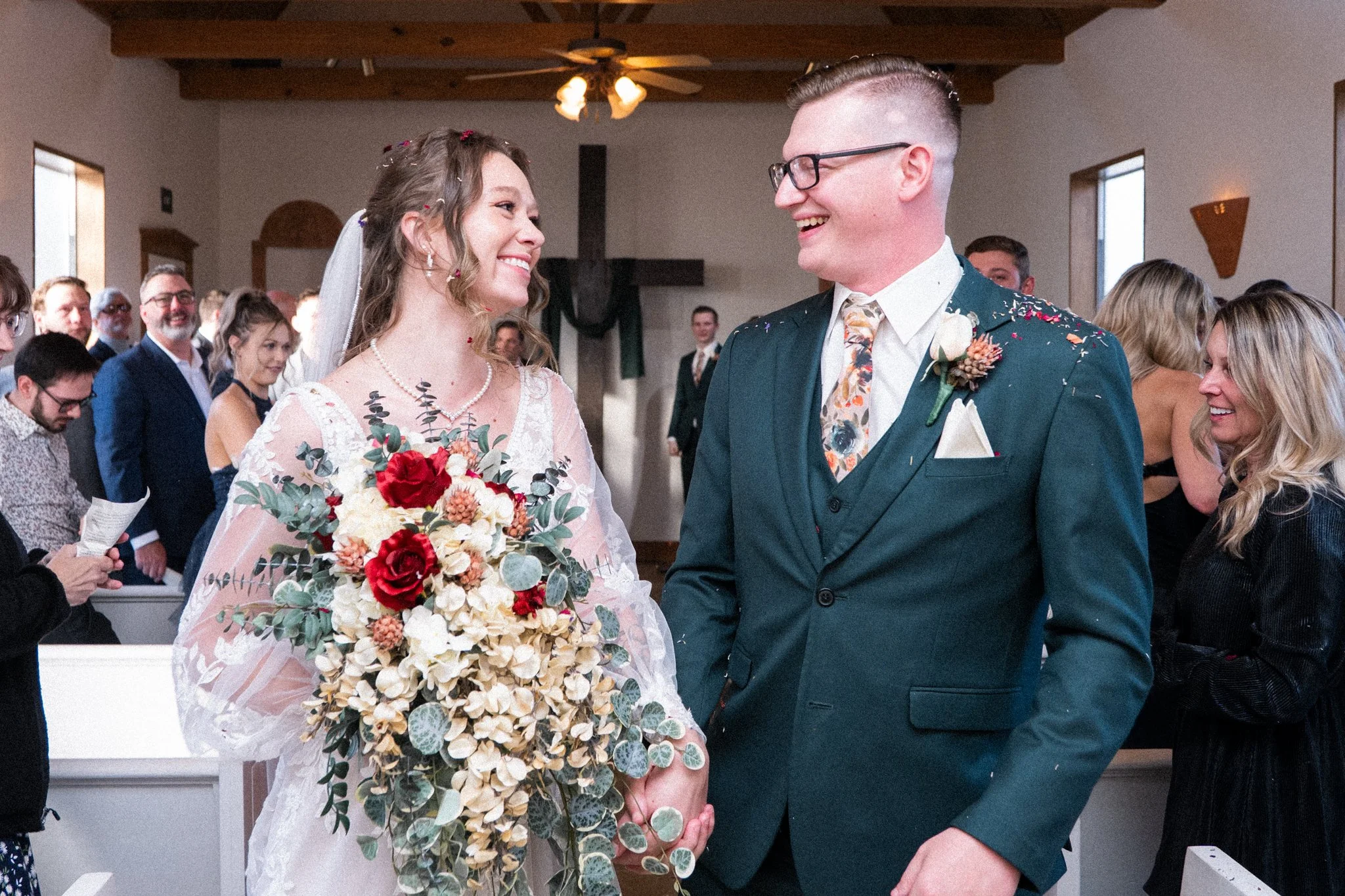 A Dallas bride and groom smiling at each other during their wedding ceremony, holding hands. The bride holds a bouquet of flowers, and guests are in the background.
