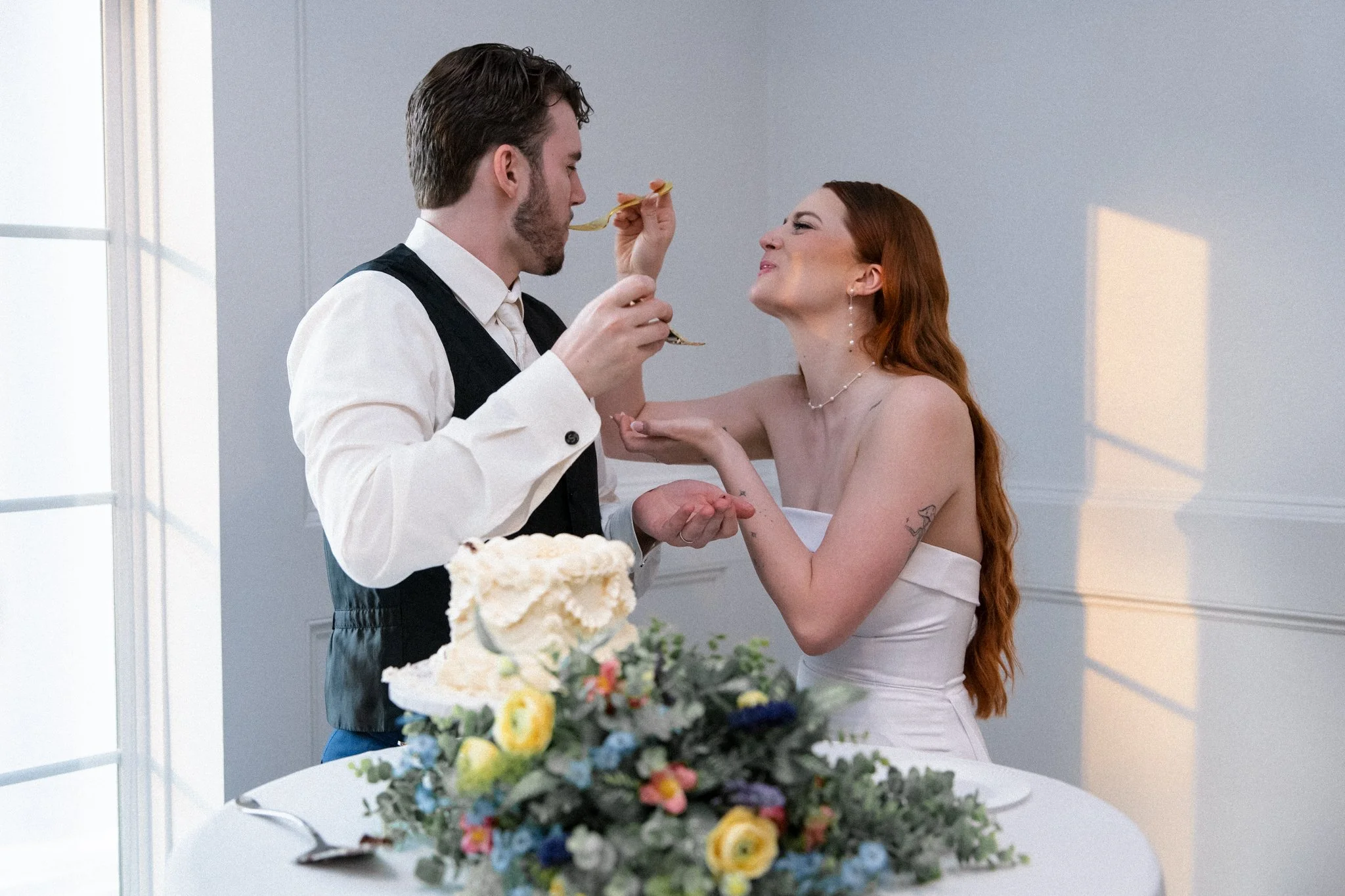 A DFW groom and bride having a playful moment during a wedding celebration, with the man feeding the woman with a fork, both smiling, in a bright room with a floral centerpiece in the foreground.