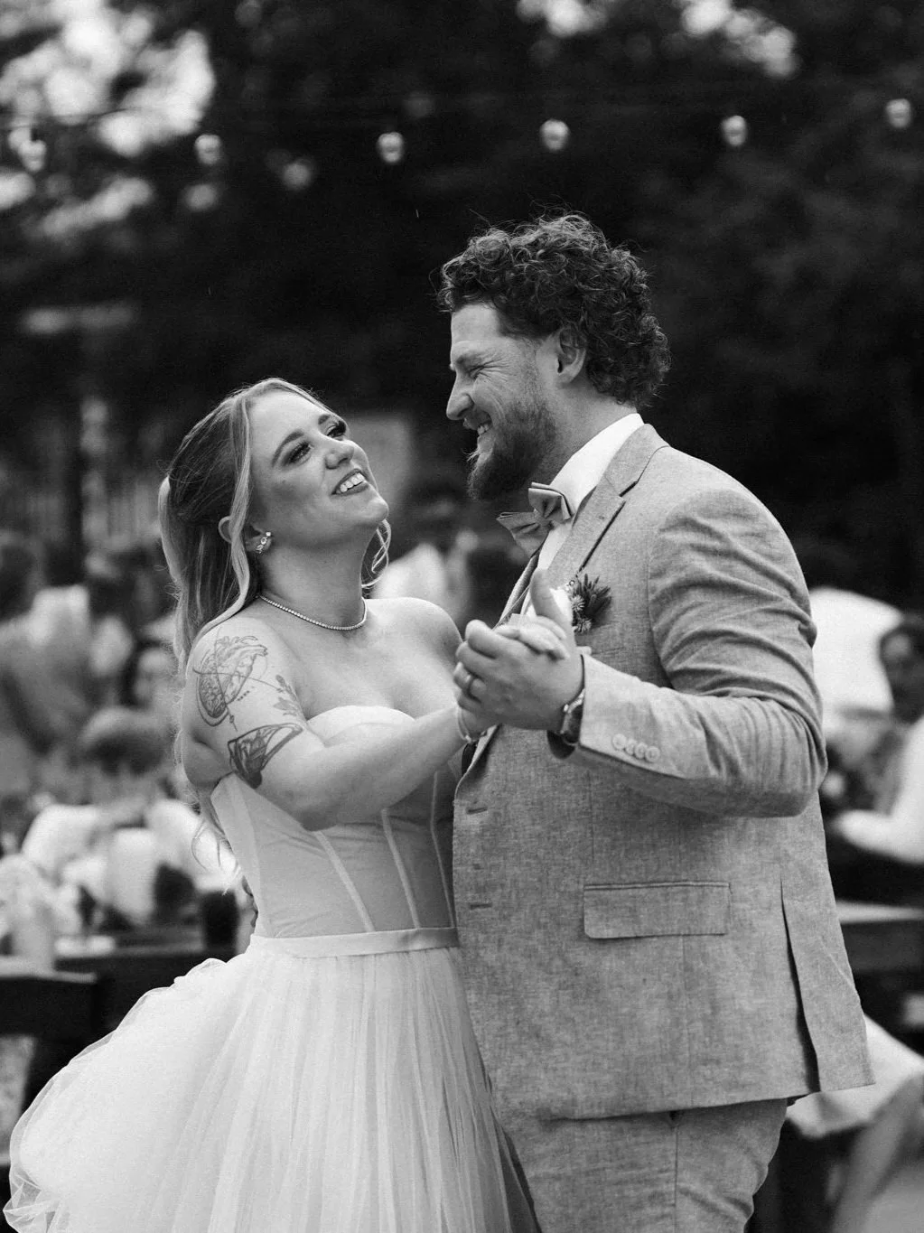 A Dallas bride and groom are dancing and smiling at their wedding, with blurred guests and string lights in the background.