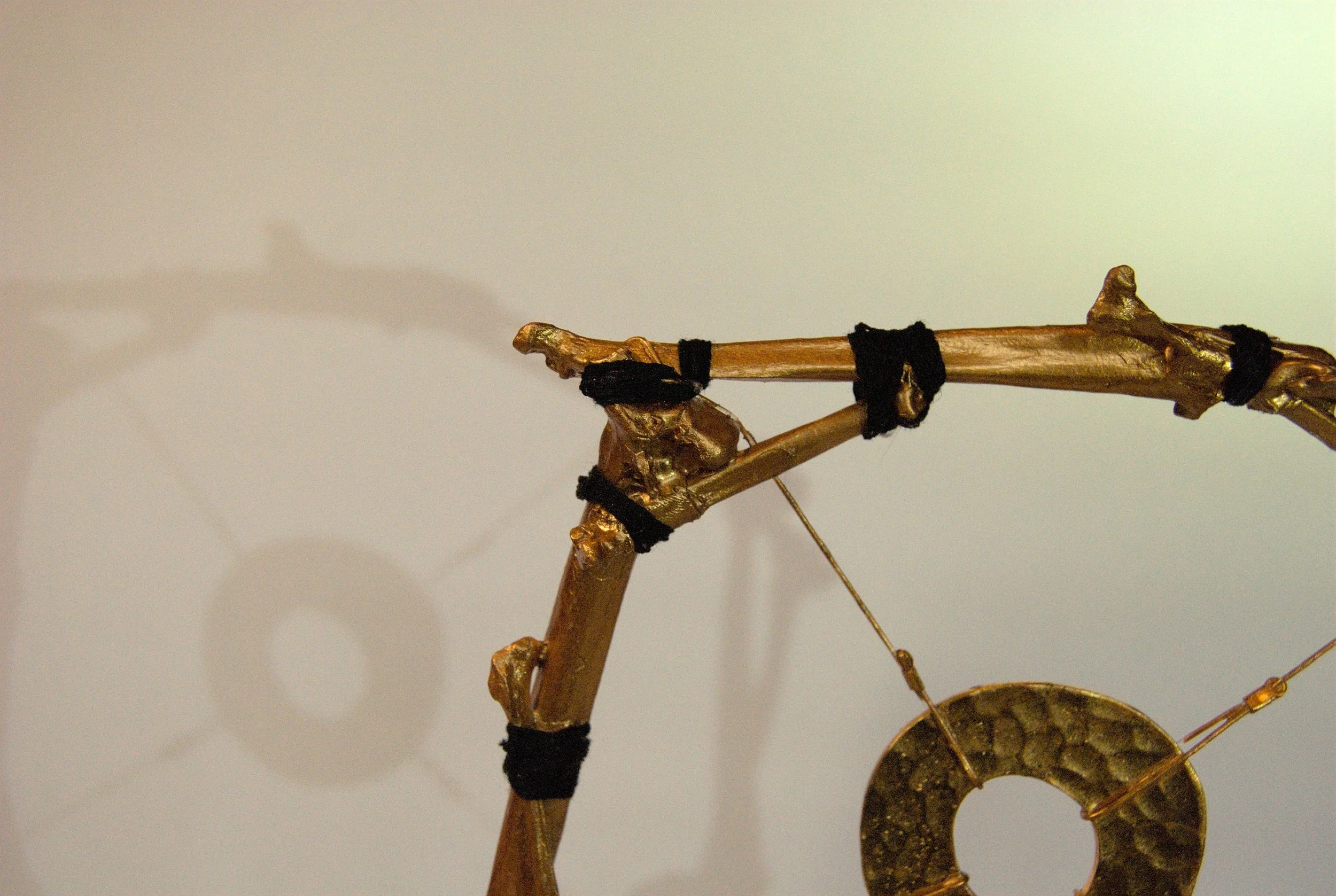 Close-up of a gold, abstract sculpture made of tiny natural bones with black fabric bindings, casting a shadow on the plain background.