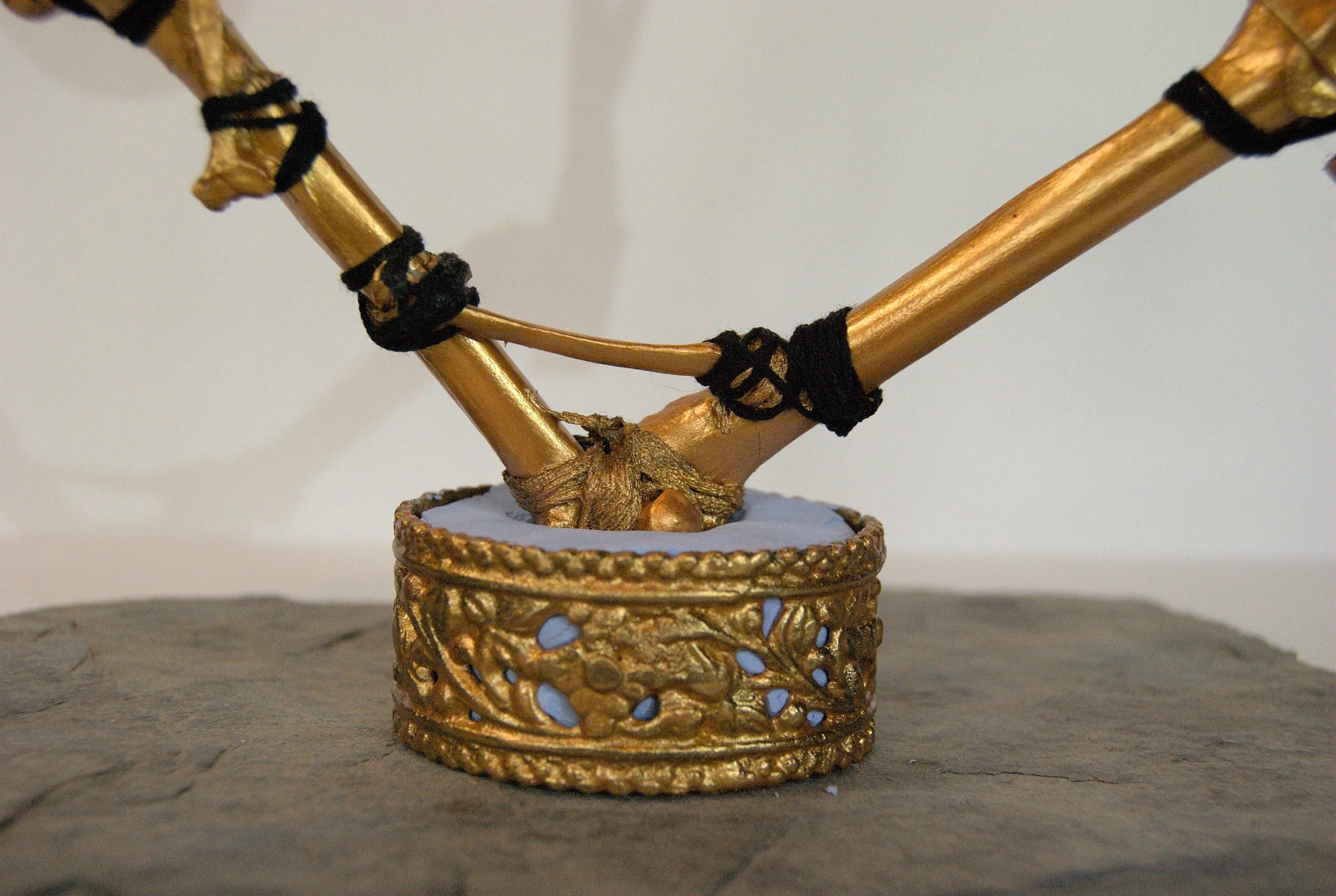 Close-up of a decorative gold-colored tree sculpture with black ties, set in a gold ornate container on a gray surface with a plain background.