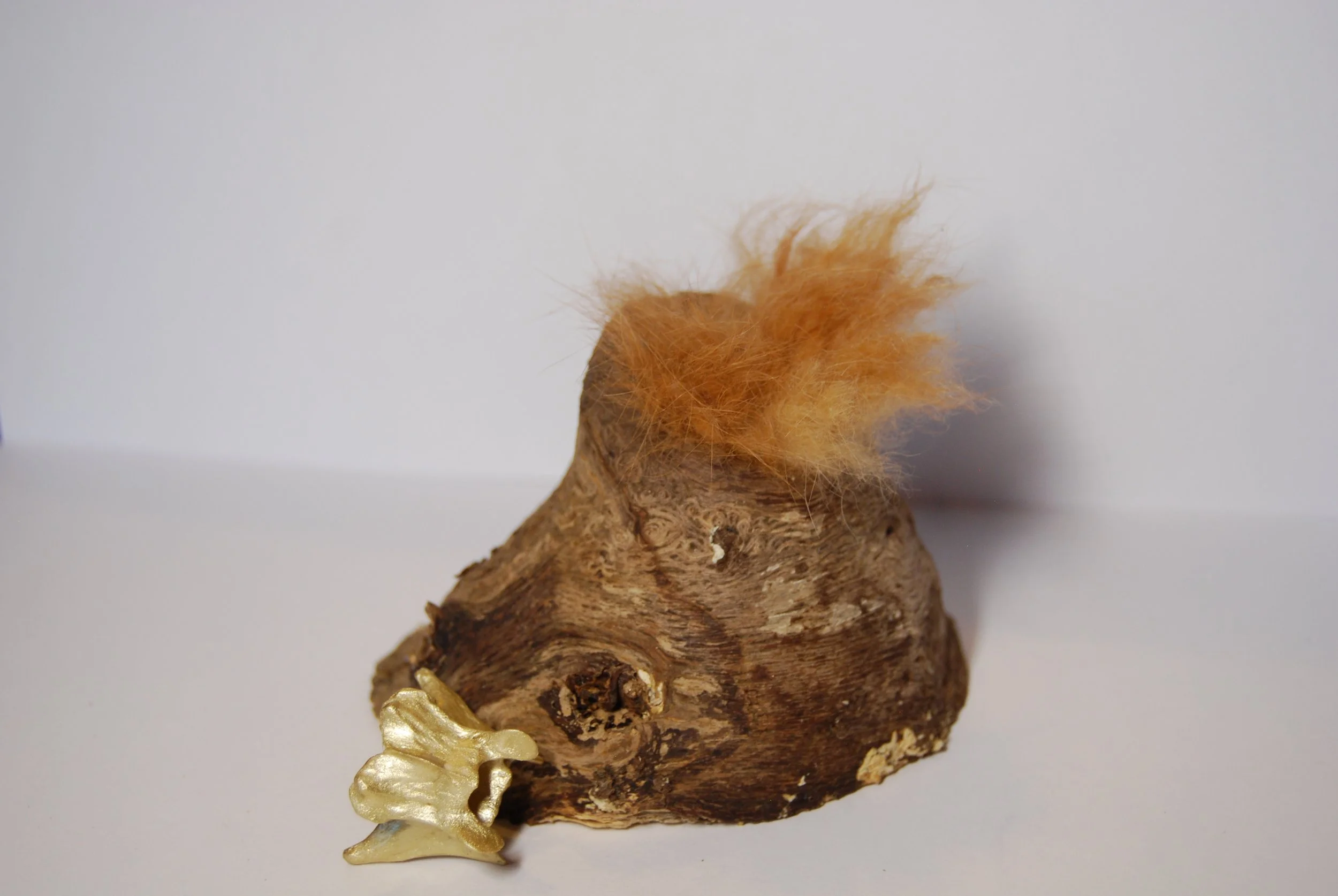 A piece of wood with orange felted hair and a metal bird figurine attached, set against a plain white background.