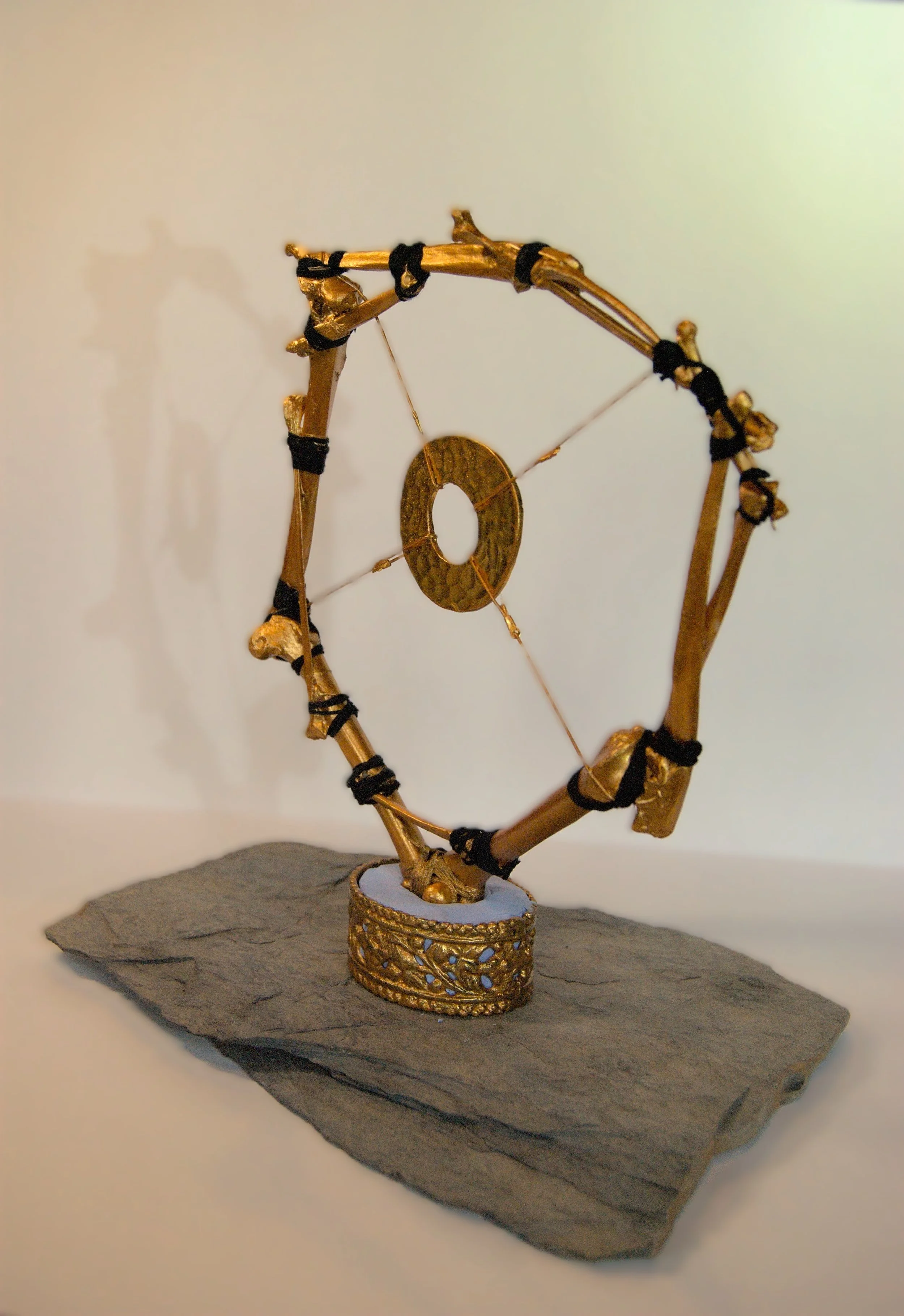 A decorative sculpture made of gold-colored bones and black ties, shaped into a circular design elevated on a dark flat stone base.