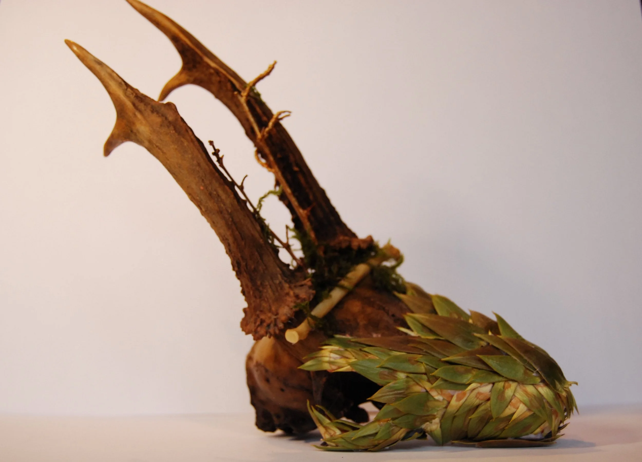 A sculpture resembling a bird, made from natural materials such as wood, moss, and leaves, positioned against a plain background.