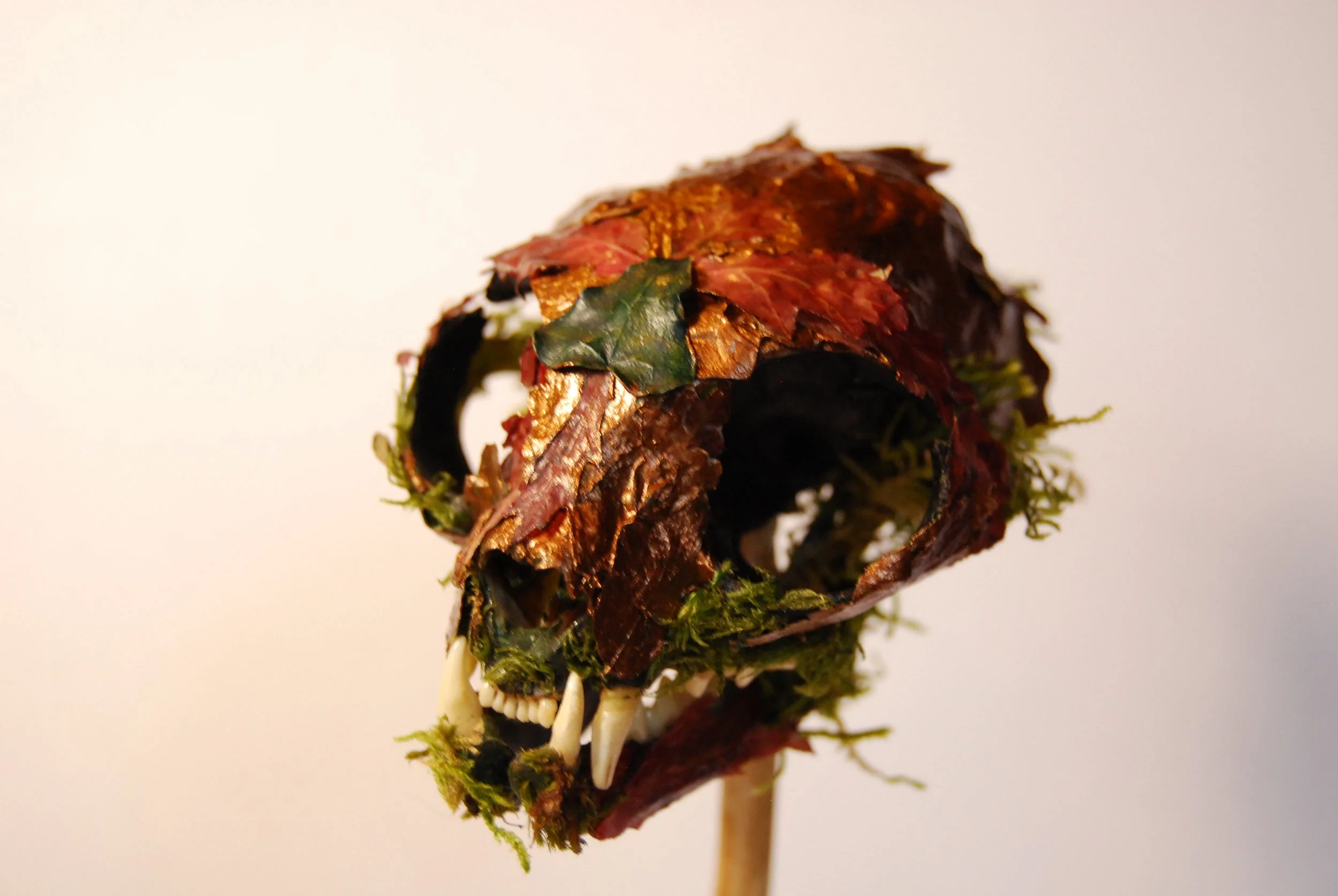 A skull mask made of leaves, moss, and animal teeth on a stick, resembling a wild animal or creature.