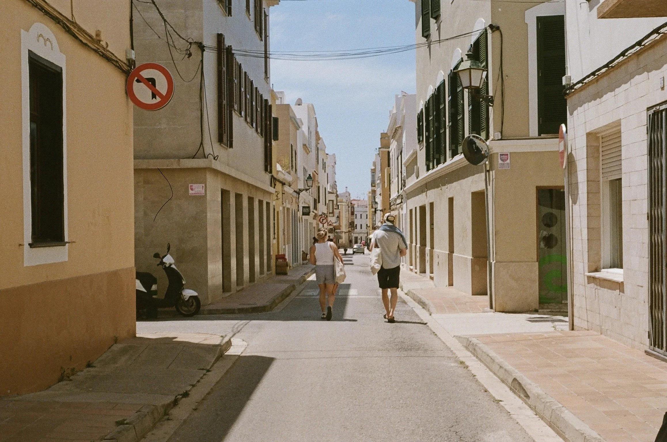 Two people walking down a narrow, sunlit street lined with pastel-colored buildings with open windows and shutters, a scooter parked on the left, and no left turn traffic sign visible. Taken in Ciutadella de Menorca, Menorca, Spain, 2024. 