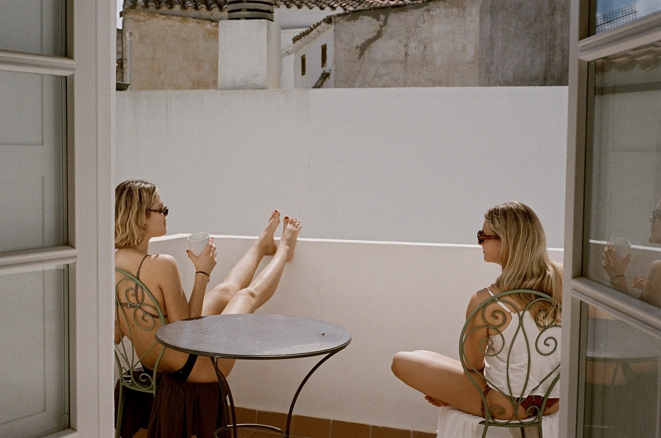 Two women sitting outside on a balcony, one with legs up on the wall, drinking from a mug, and the other sitting with legs crossed, both wearing sunglasses and casual summer clothing. Taken in Ciutadella de Menorca, Menorca, Spain, 2024.