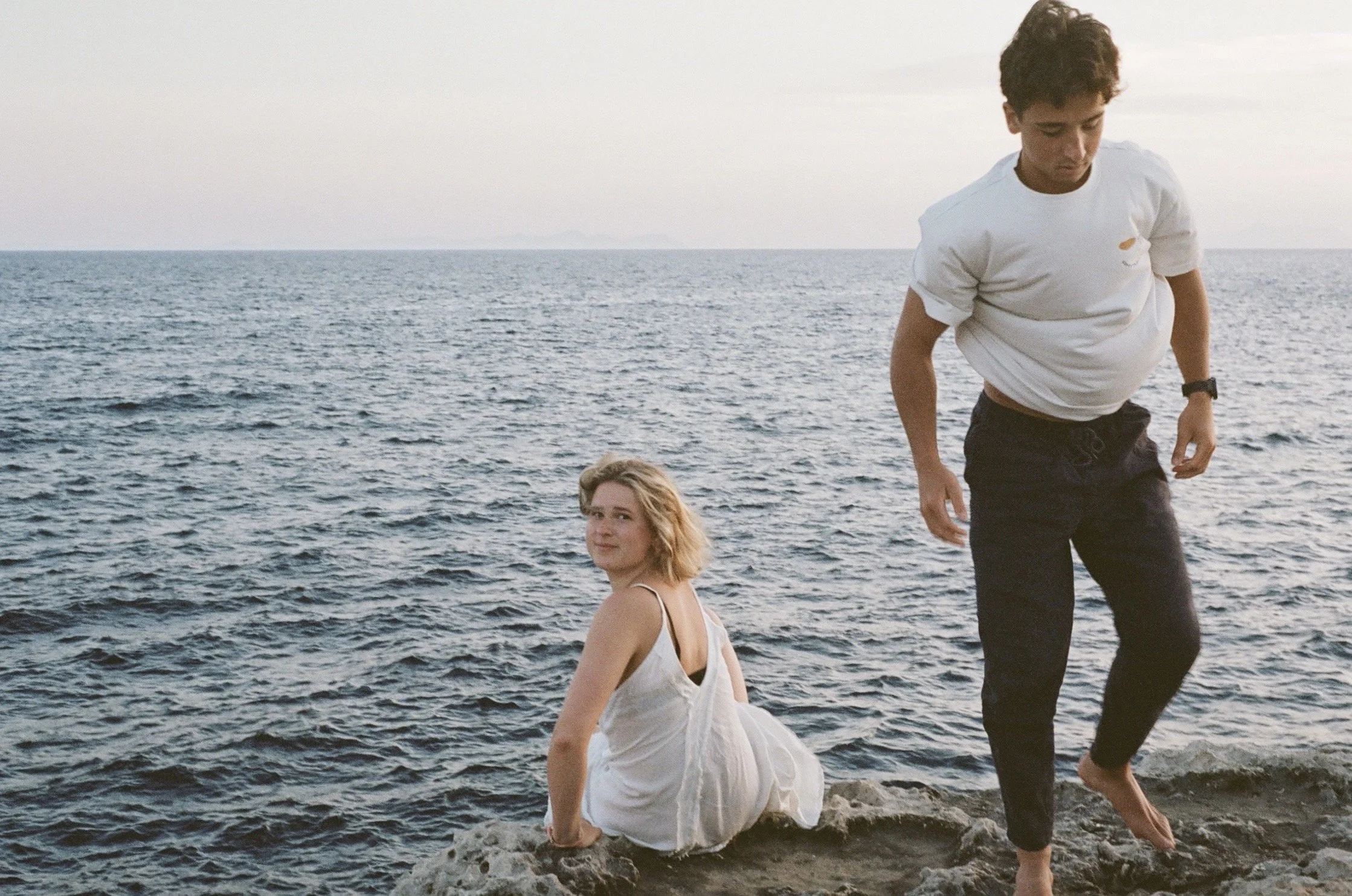 A young woman with blonde hair, sitting on rocks by the water, looking at the camera. A young man with dark curly hair, walking barefoot on the rocks, looking down. They are near the ocean with a calm sea and a cloudy sky.