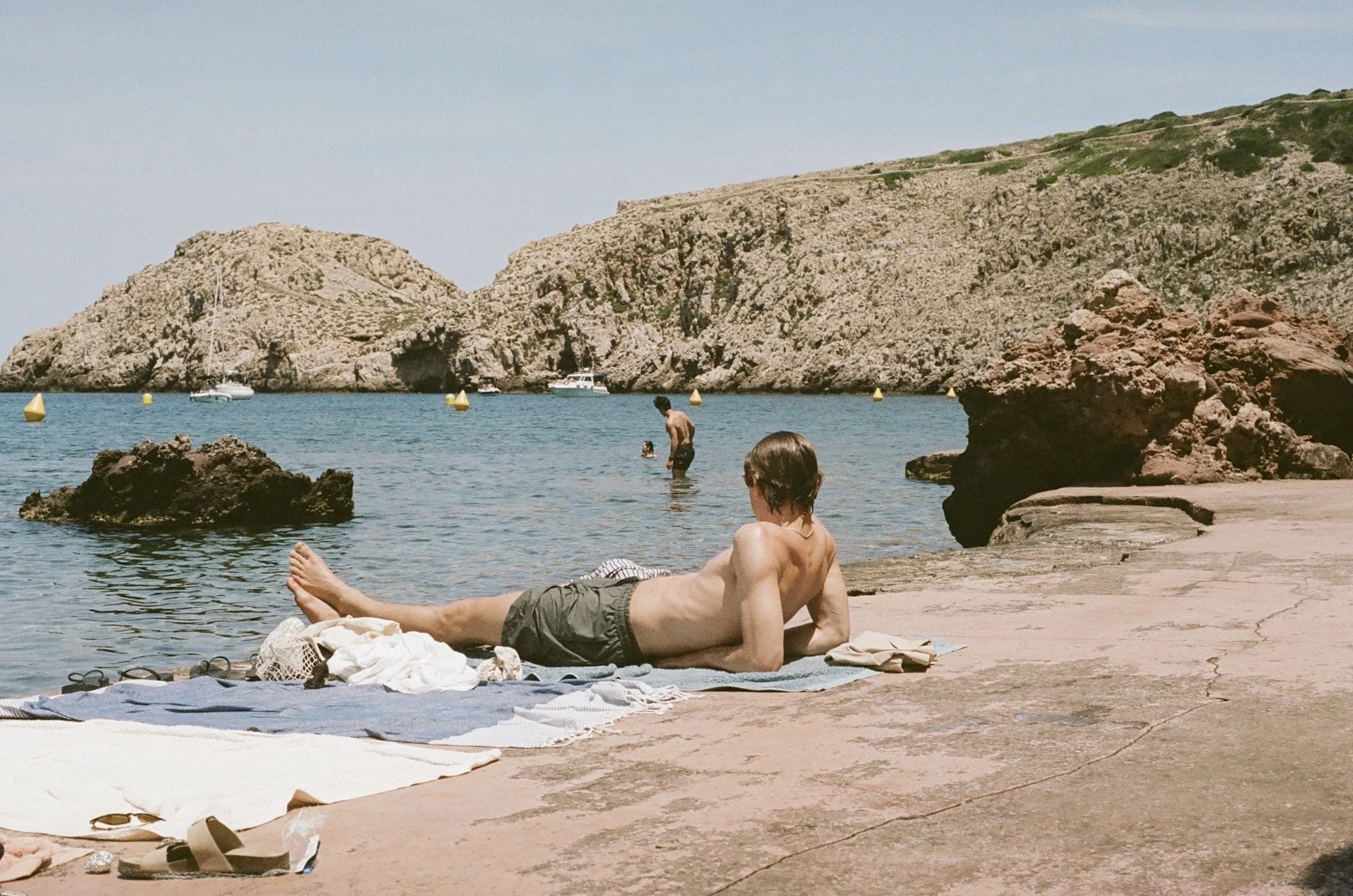 A man relaxing on a towel on a rocky beach with others swimming and boats in the water, rocky cliffs in the background. Taken at Cala Morell, Menorca, Spain, 2024.