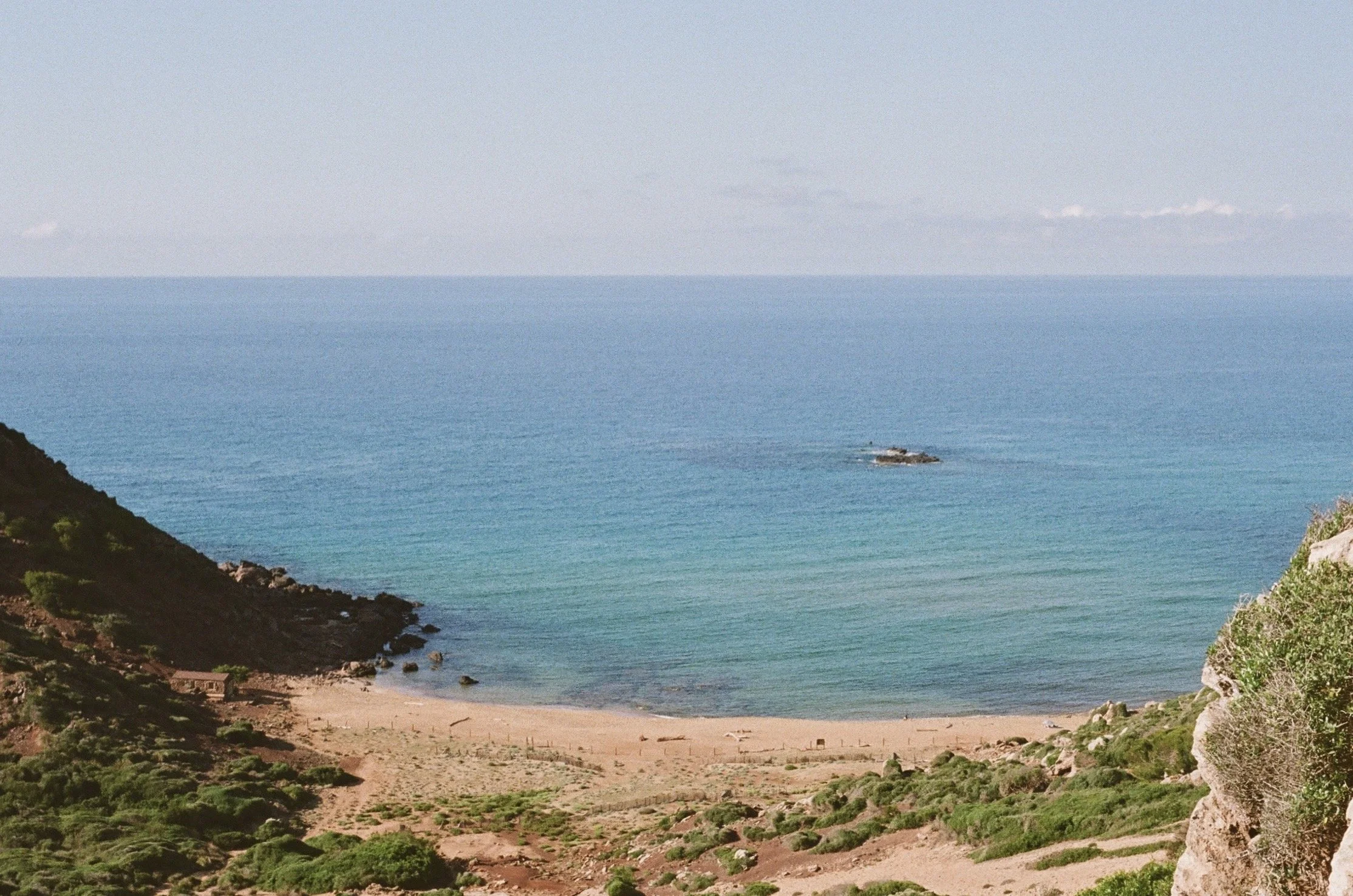 A coastal view with a sandy beach between rocky hills, calm blue ocean waters, and a small island in the distance under a clear sky. View of Cala del Pilar, Menorca, Spain, 2024. 