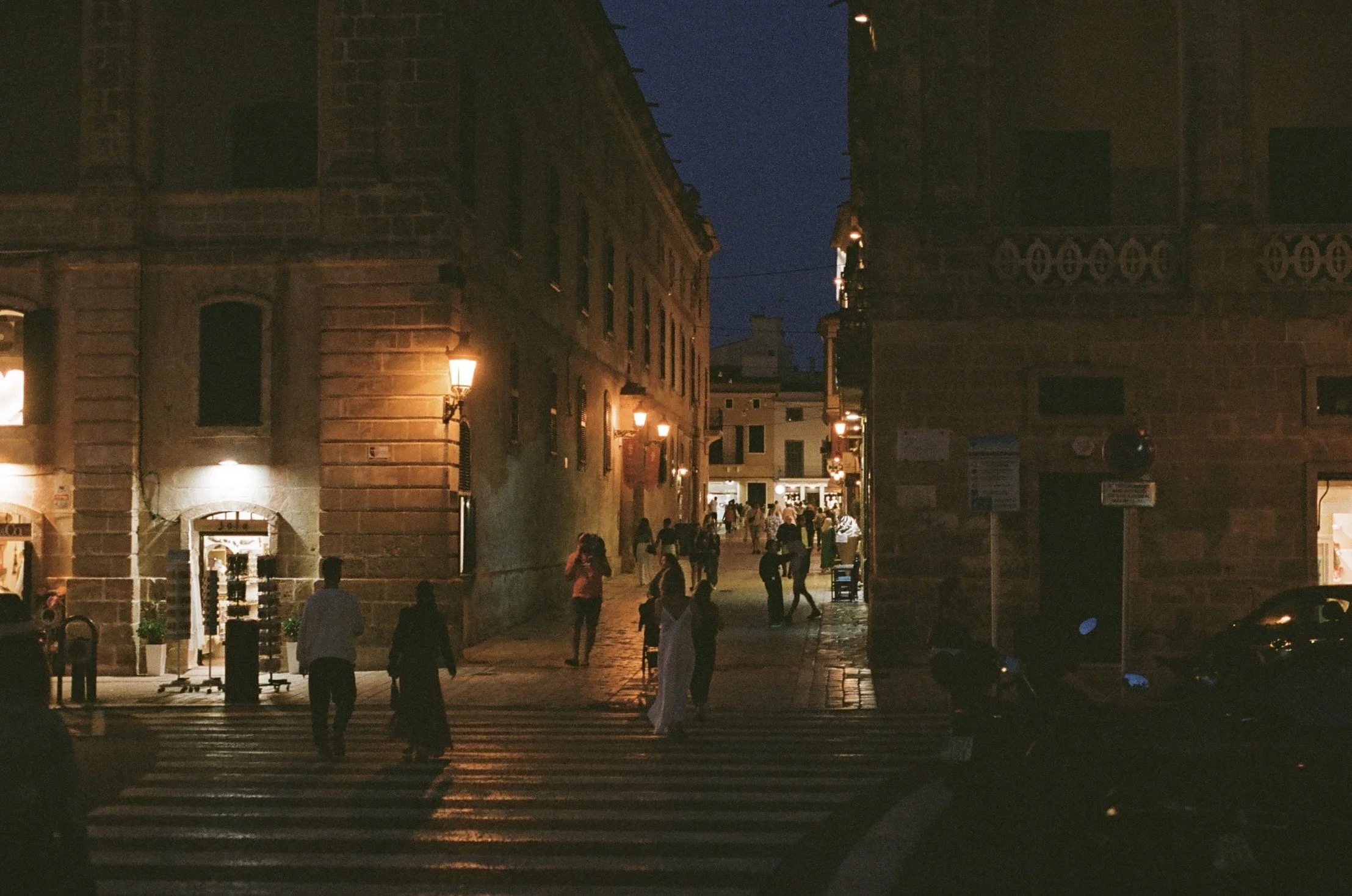 A nighttime street scene in a historic city, with pedestrians walking on a crosswalk and streetlights illuminating brick buildings on both sides. Taken in Ciutadella de Menorca, Menorca, Spain, 2024. 