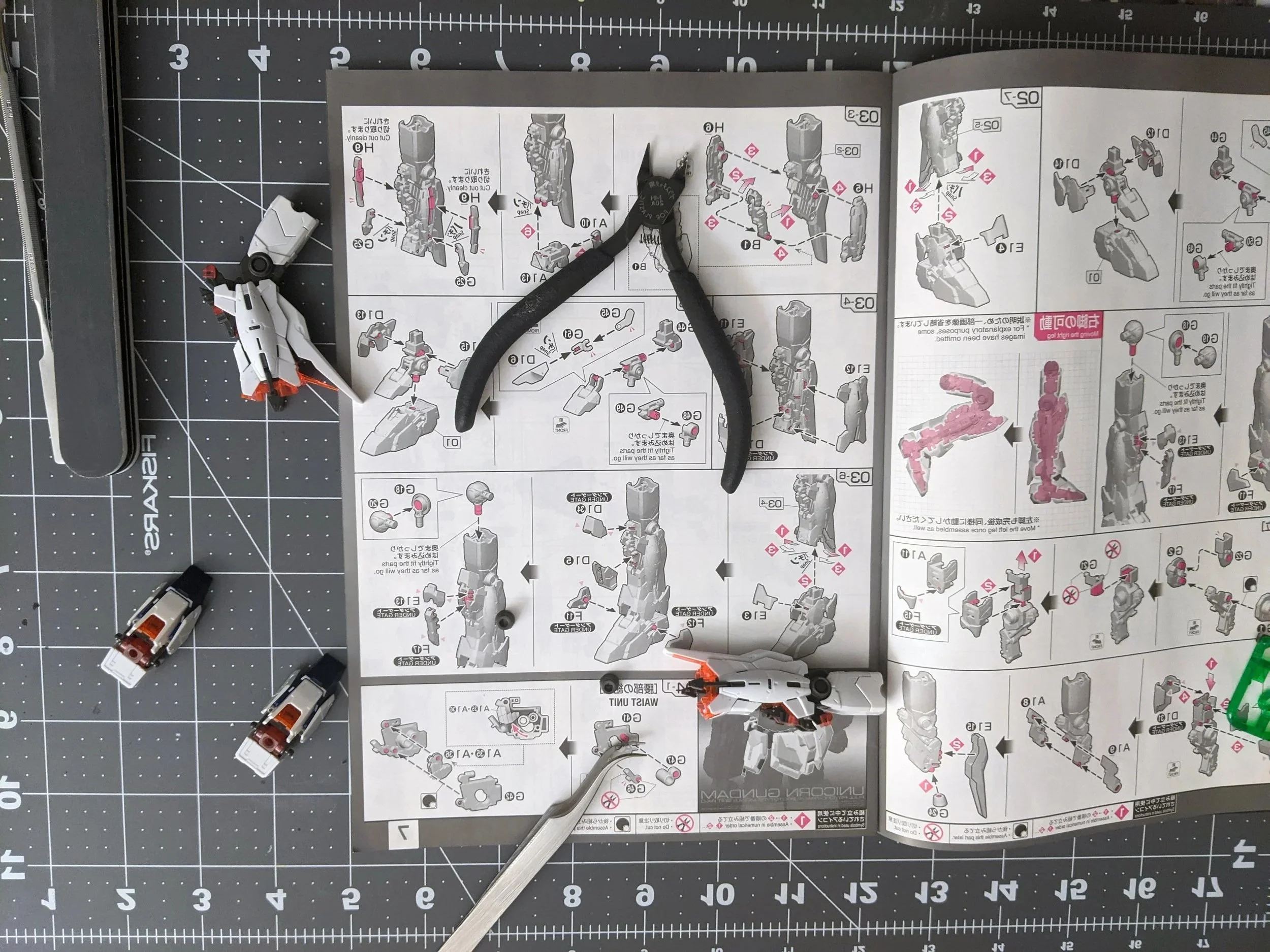 Overhead view of a work surface with an assembled plastic model kit, instruction manual, pliers, a metallic hobby knife, and small plastic parts, all arranged on a cutting mat with measurement markings.