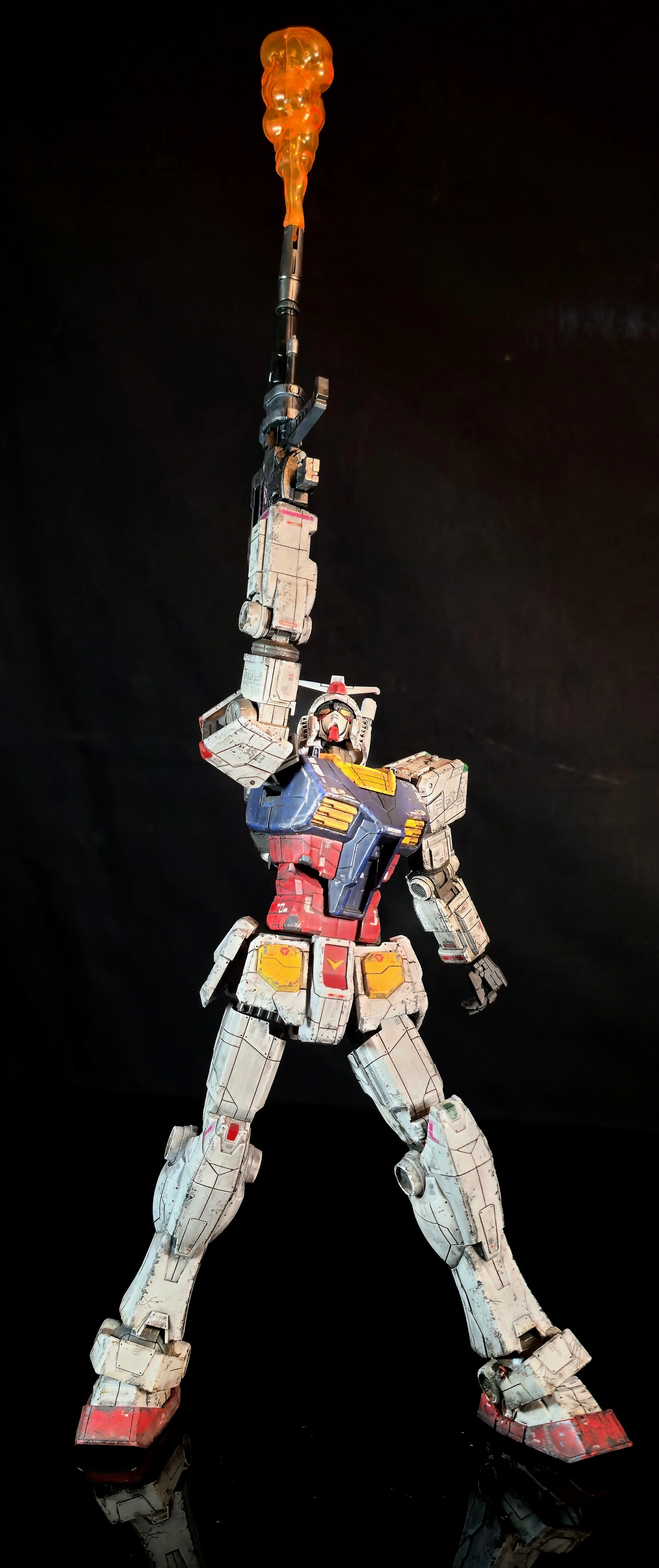 A model of a Gundam robot figure holding a glowing fiery orange weapon high above its head, set against a black background.