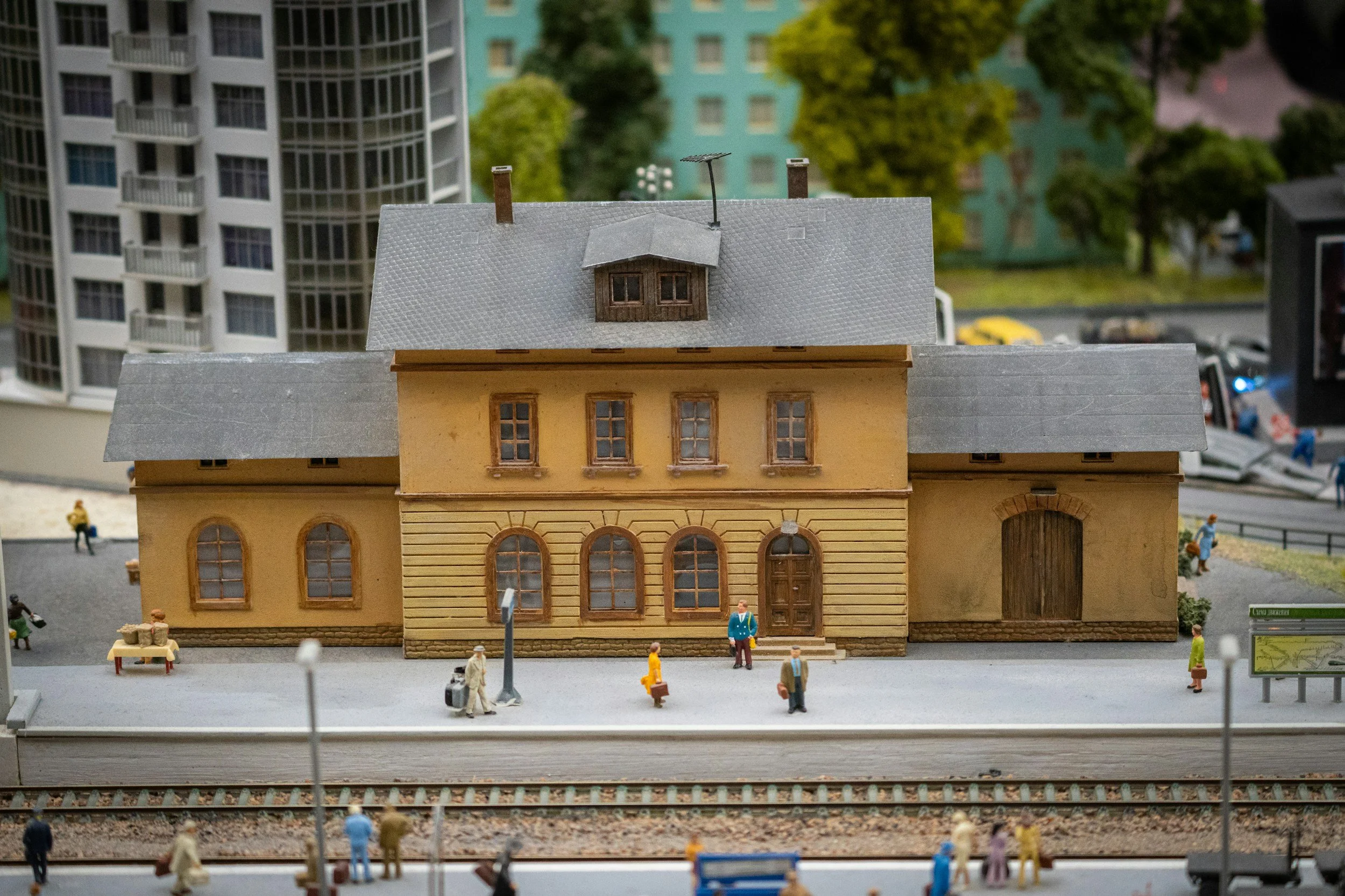 Close-up of a detailed miniature model of a yellow building with grey roof, small windows, and miniature figures around it, set against a backdrop of other buildings and trees.