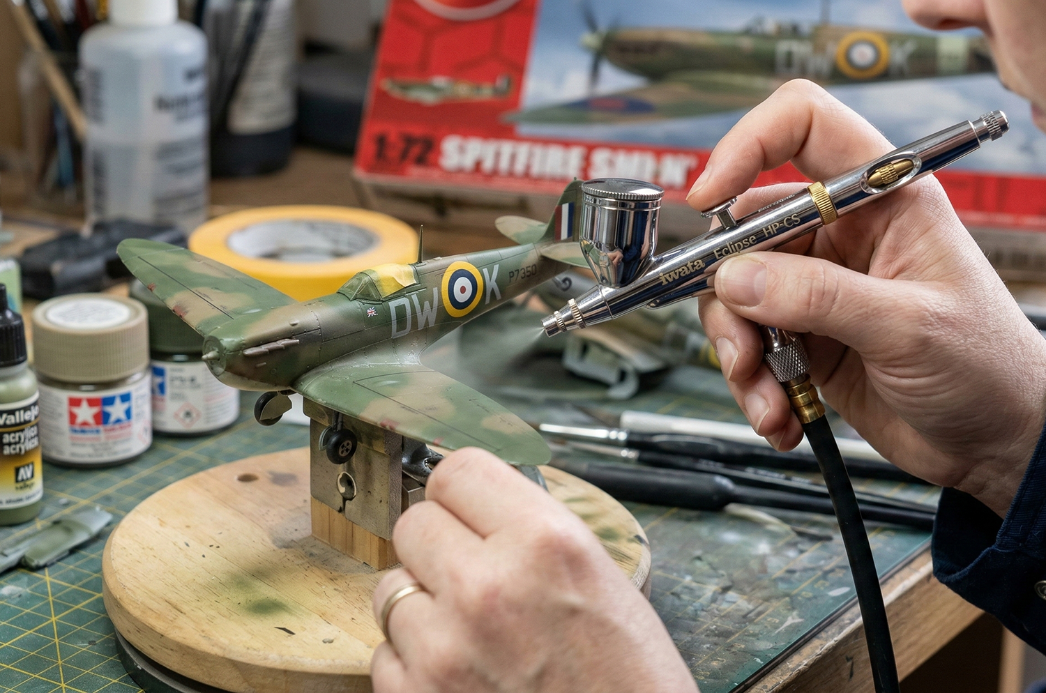 Person working on a detailed model airplane, using an airbrush to paint a camouflage pattern.