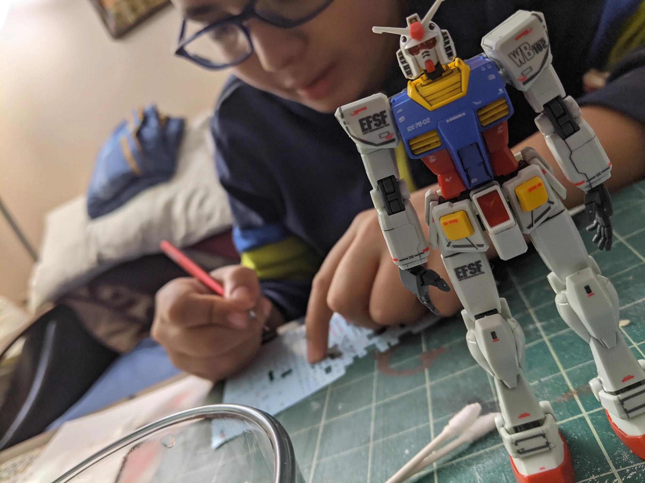 A person with glasses and a dark shirt is sitting at a table, writing on paper, with a model robot figure in the foreground.
