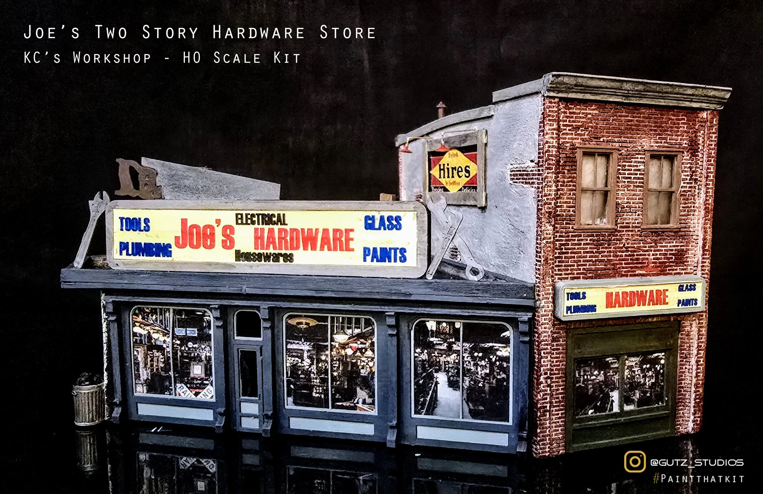 A detailed miniature model of Joe's Two Story Hardware Store and KC's Workshop - HO Scale Kit, featuring a brick and gray storefront with large windows and a sign displaying tools, plumbing, electrical, glass, paint, and hardware. The background is black, and there are vintage signs on the building, with reflections visible in the windows.