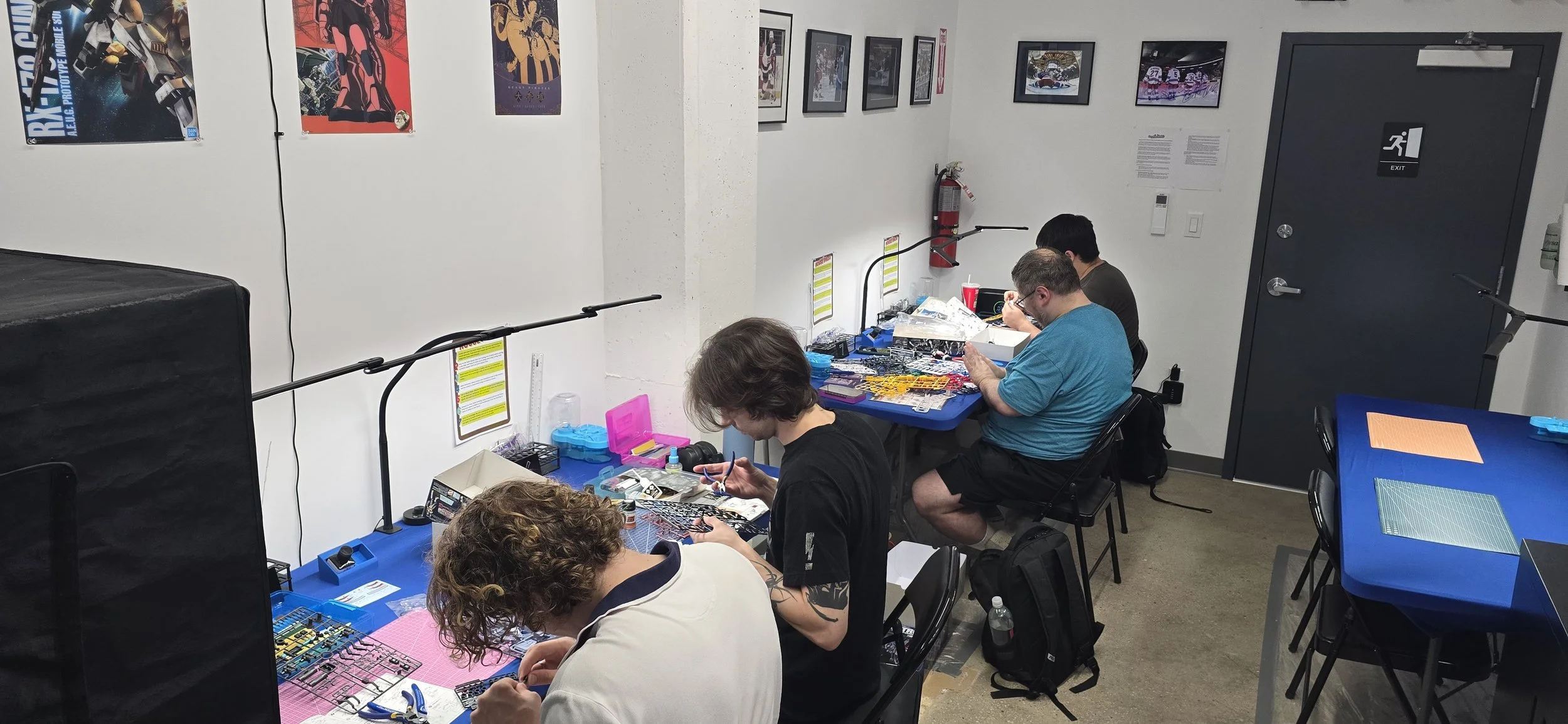 Guest from out Gunpla and Chill Thursday Nights working at tables with plastic model kits and hobby tools in a workshop setting, with framed artwork on the walls and a fire extinguisher near the door.