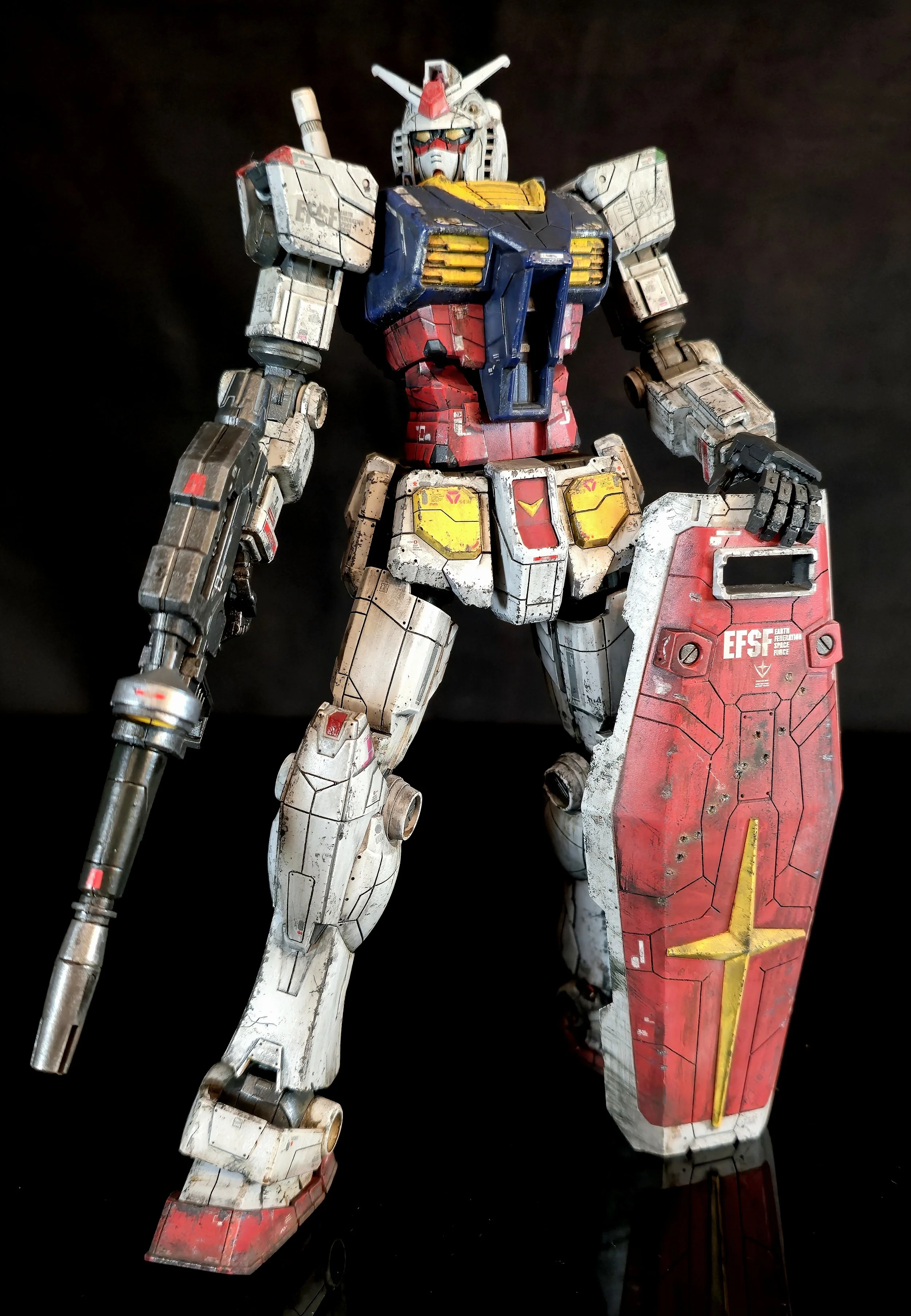 A detailed model of the Gundam RX-78-2 in white, blue, red, and yellow, standing on a reflective surface against a black background.